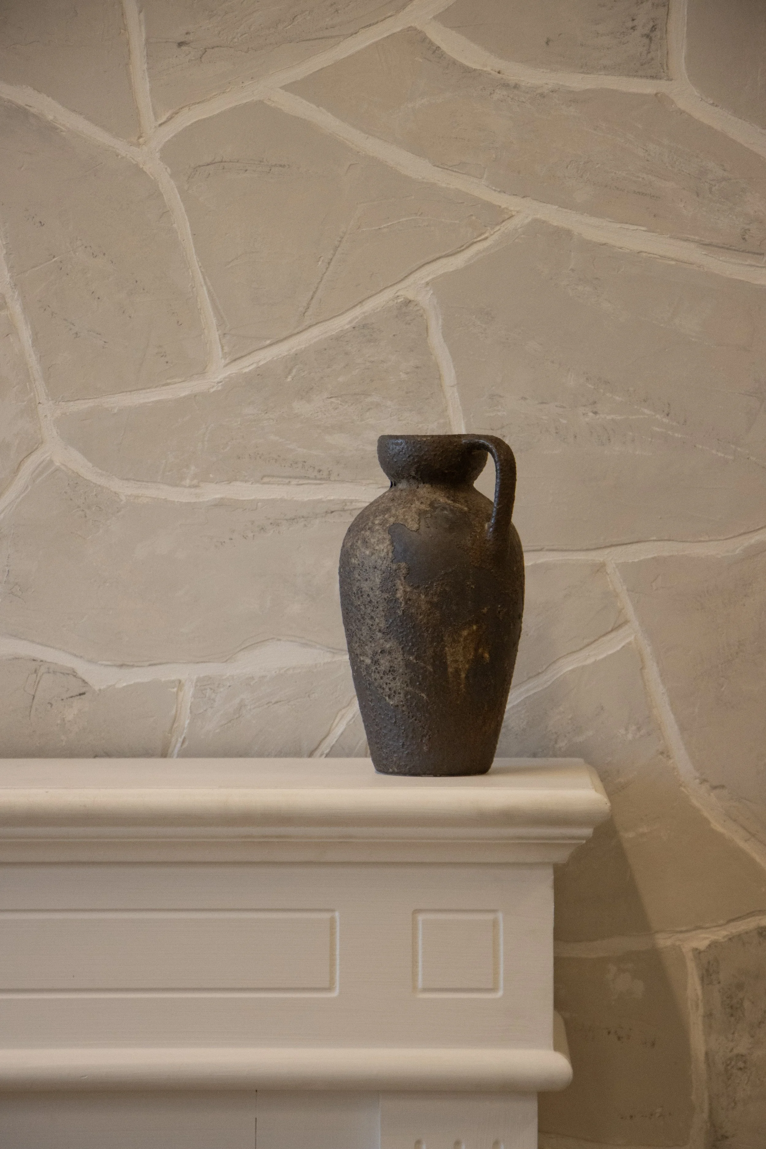 A dark, weathered ceramic vase with a small handle on one side, placed on a white shelf against a beige stone wall.