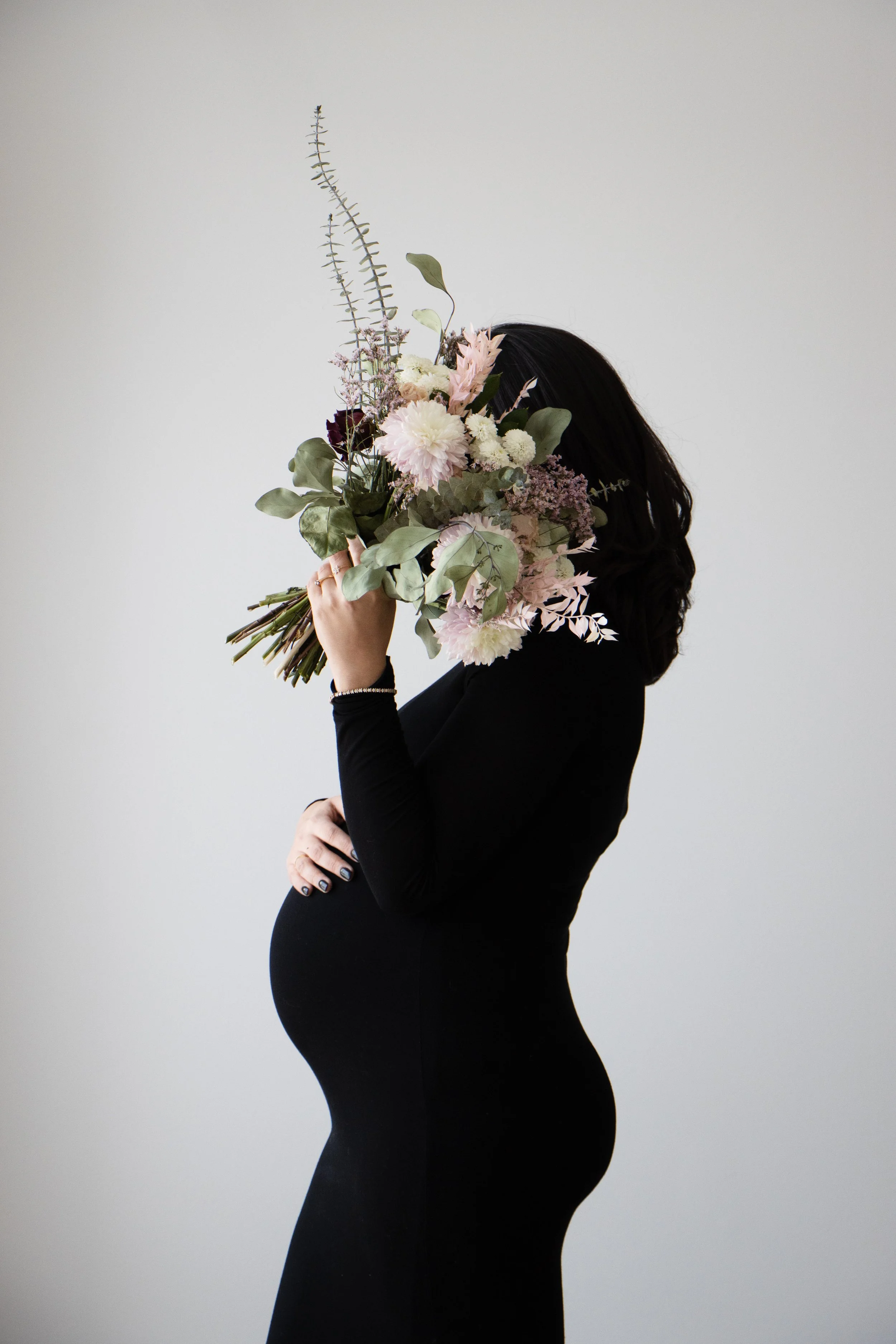 A pregnant woman in a black dress holding a large bouquet of flowers covering her face, standing against a plain white background.