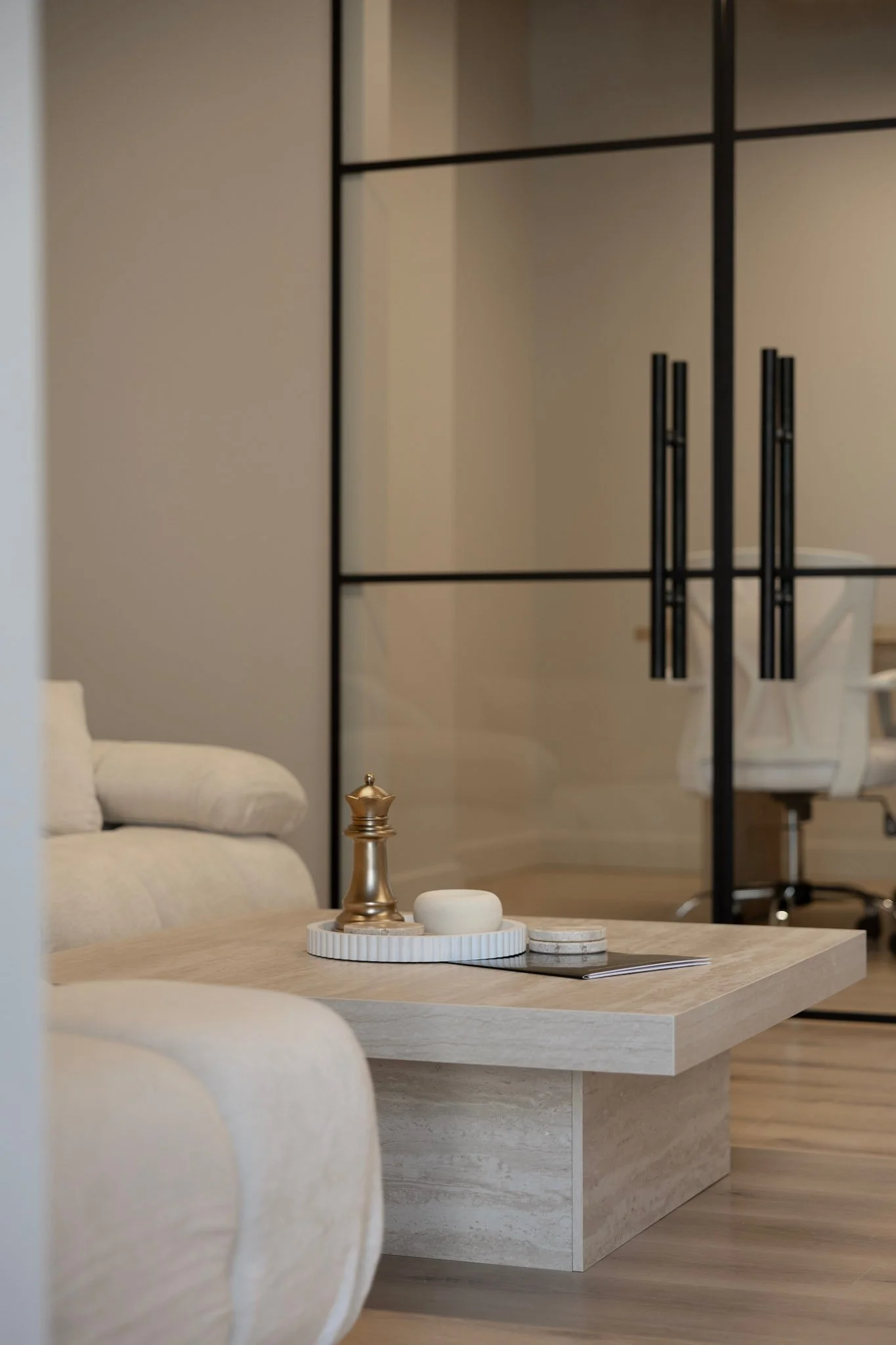 Modern living room with a beige sofa, a minimalist wooden coffee table with decorative objects, and glass partitions with black frames in the background.