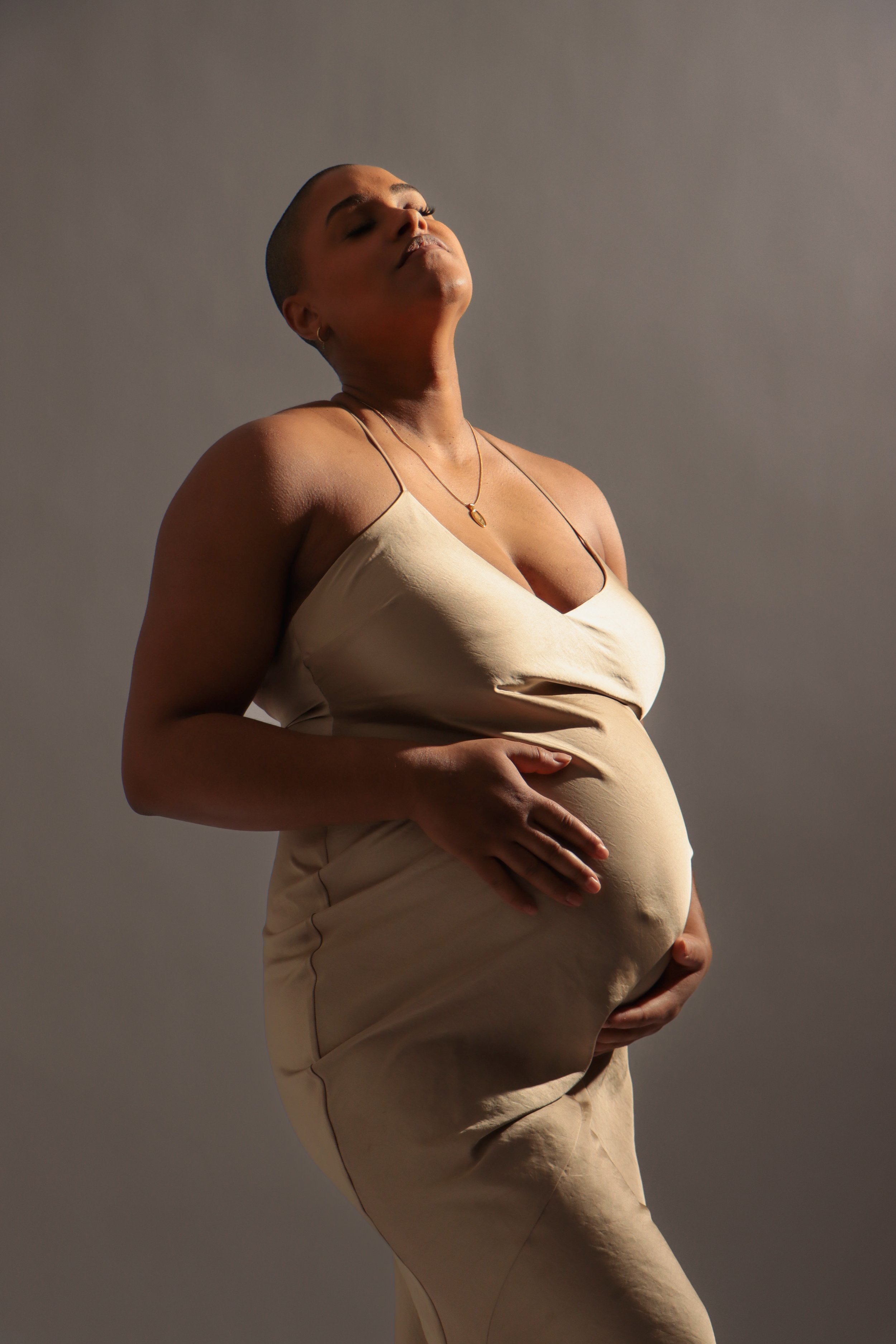 A pregnant woman with short hair, wearing a beige satin dress, gently holding her belly with both hands, with her eyes closed and head tilted slightly upwards in a serene pose.