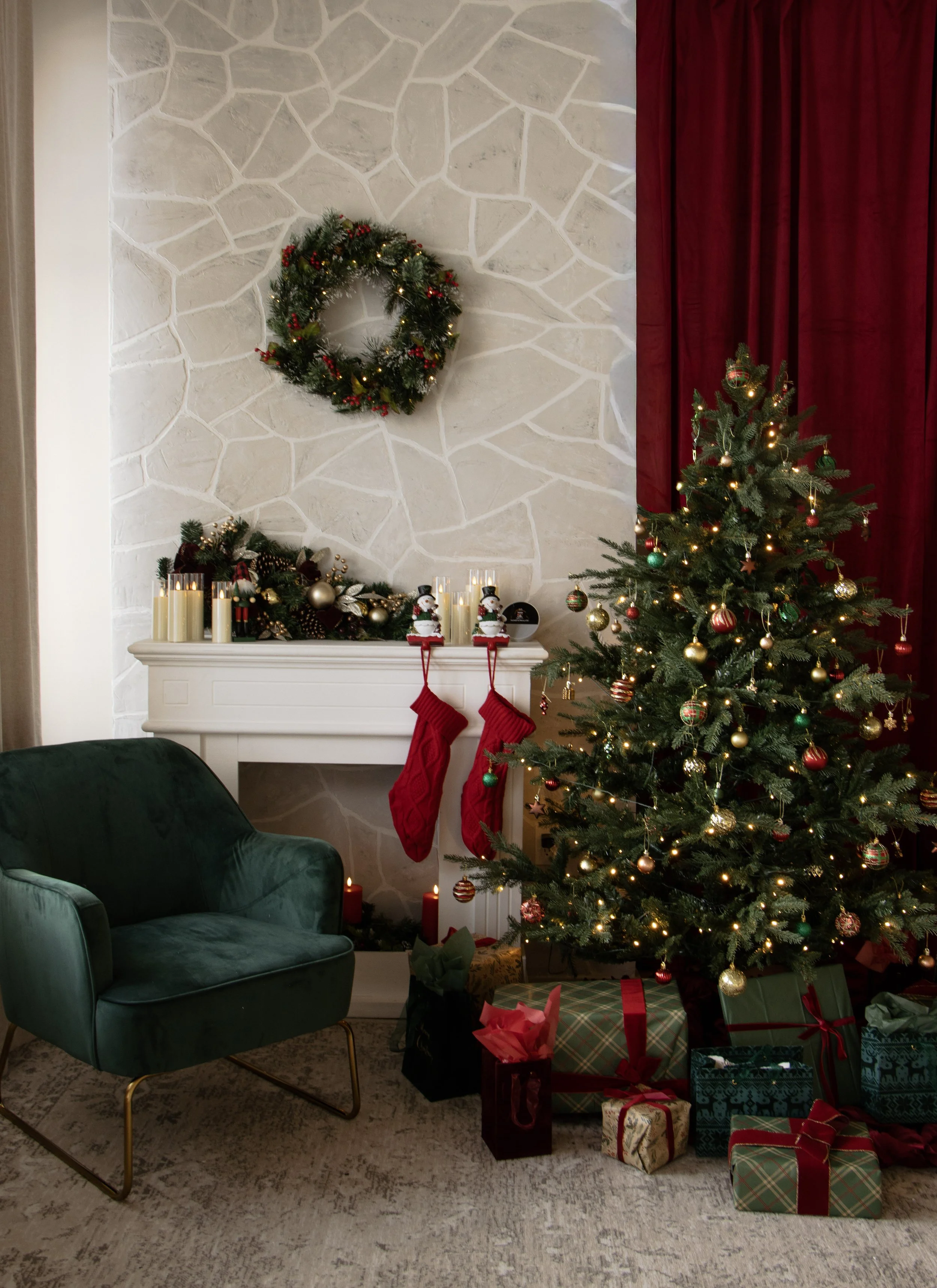 A decorated Christmas living room with a Christmas tree adorned with ornaments and lights, a fireplace with stockings and holiday decorations, a green armchair, and a pile of wrapped presents on the floor.