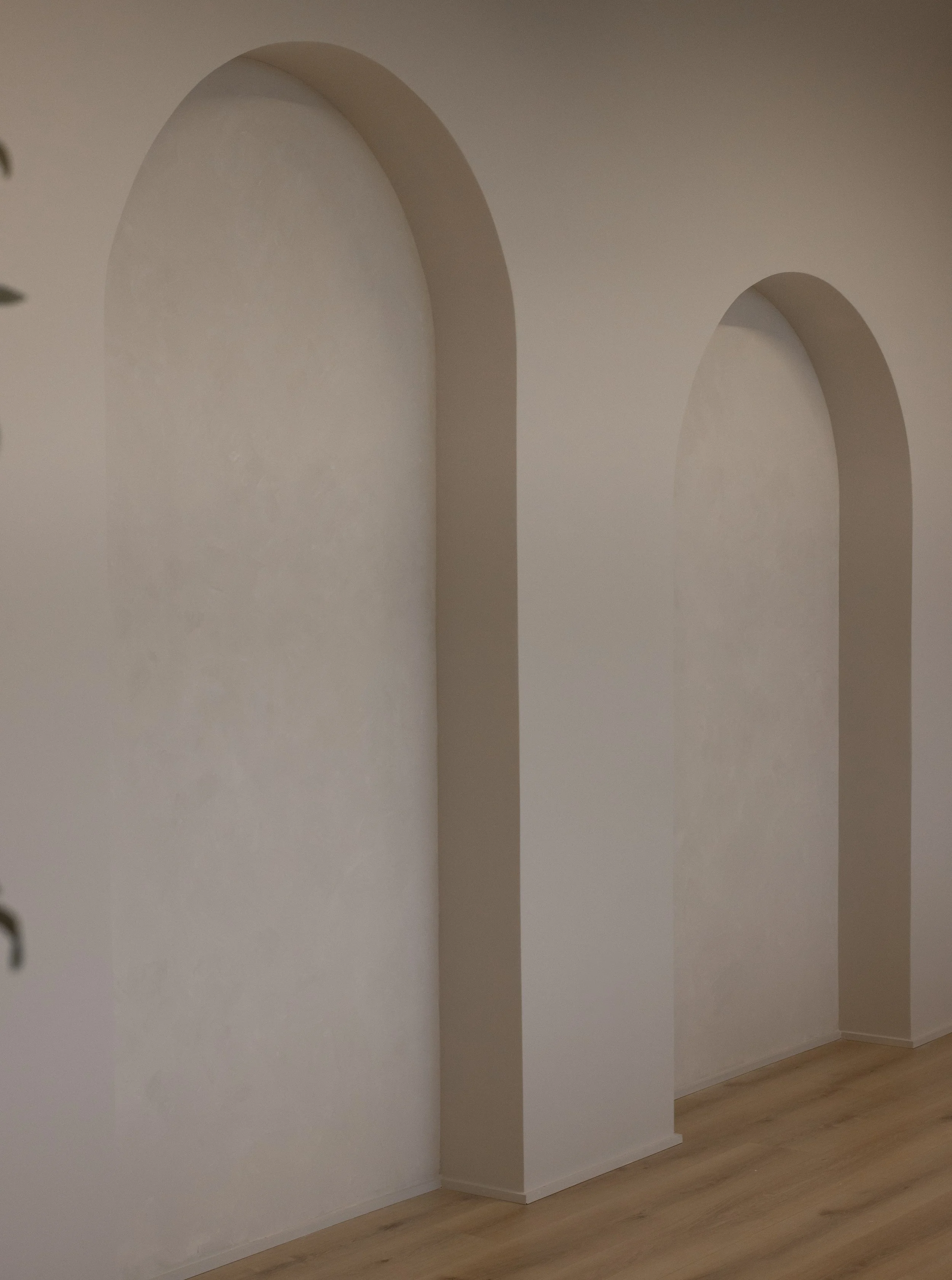 Interior wall with two arched architectural niches painted in a neutral color, next to a light wooden floor.