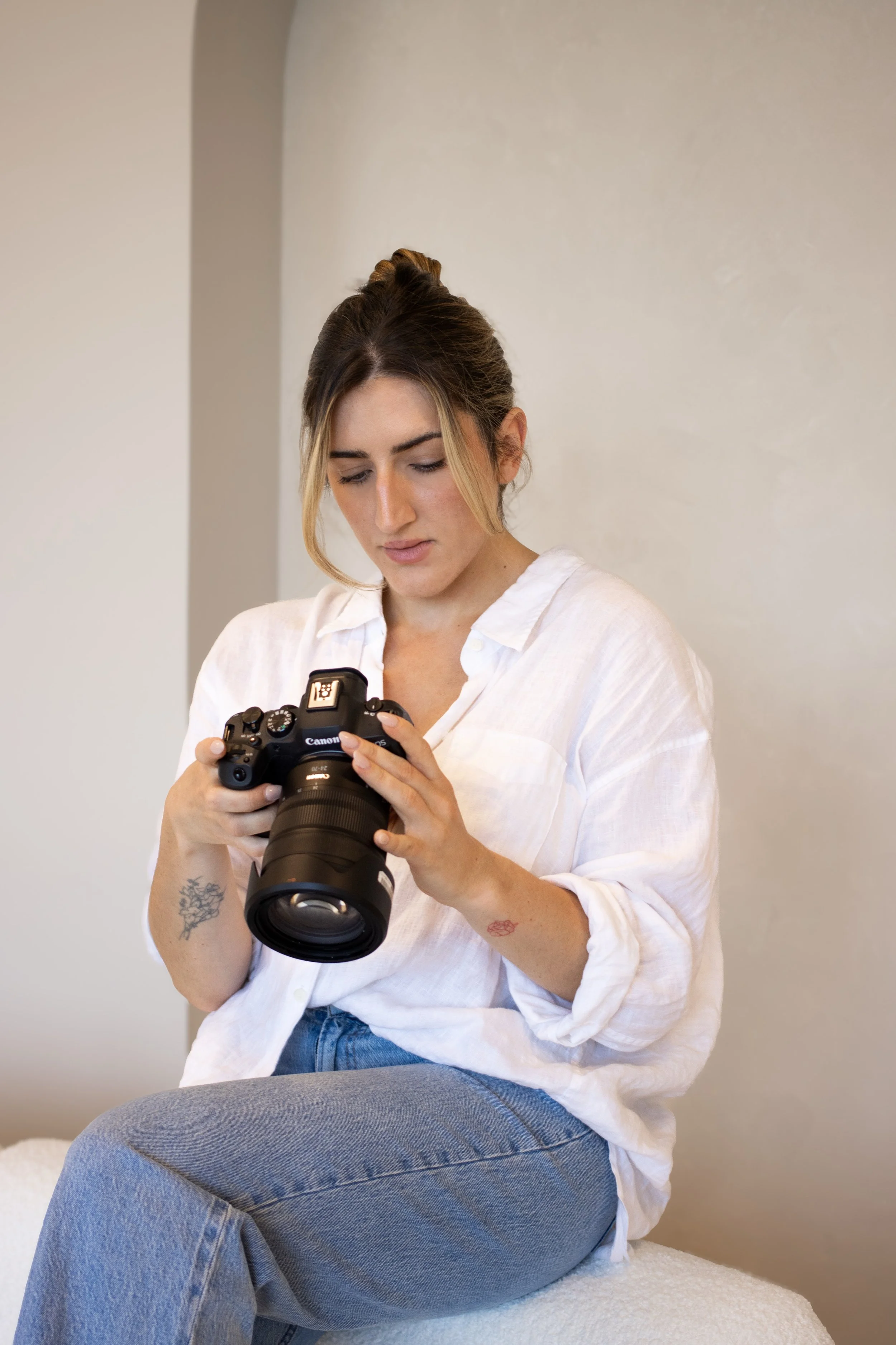 A young woman with tattoos on her right arm and left wrist, wearing a white shirt and blue jeans, sitting on a surface and looking at a professional Canon camera.