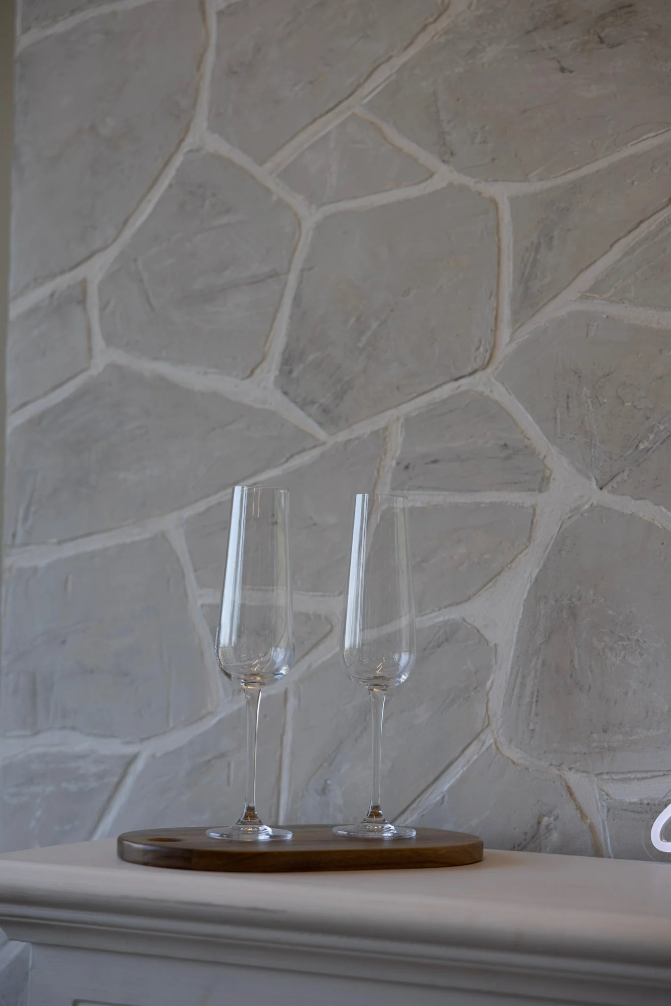 Two empty champagne flutes on a wooden tray placed on a white mantelpiece, with a stone wall in the background.