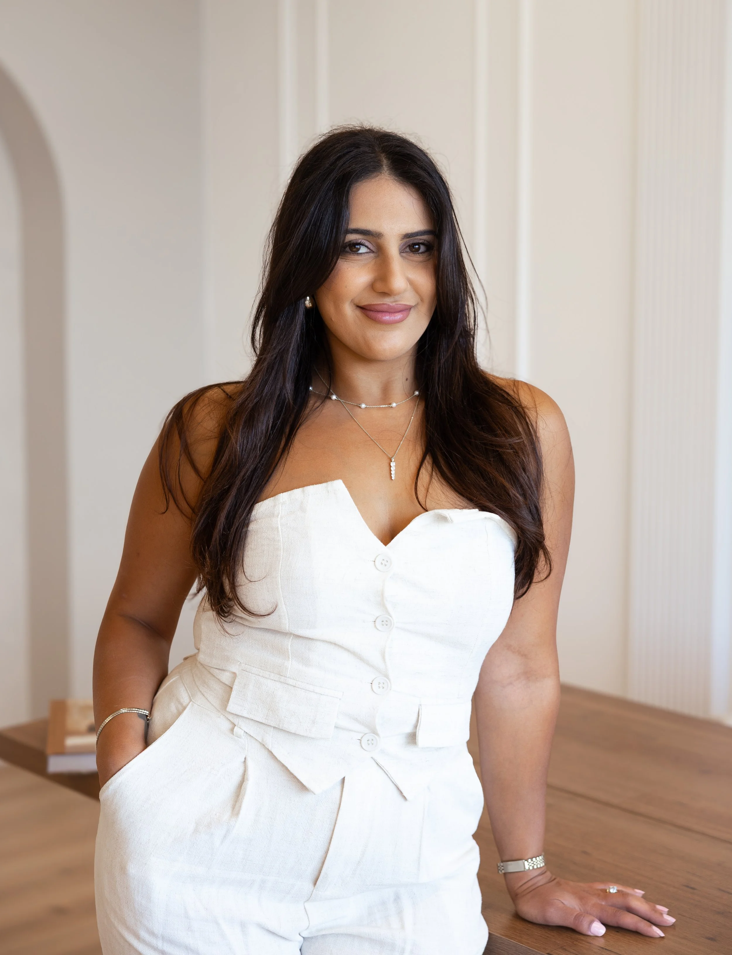 A woman wearing a white sleeveless button-up dress with her right hand resting on a wooden table, indoors with neutral-colored walls.