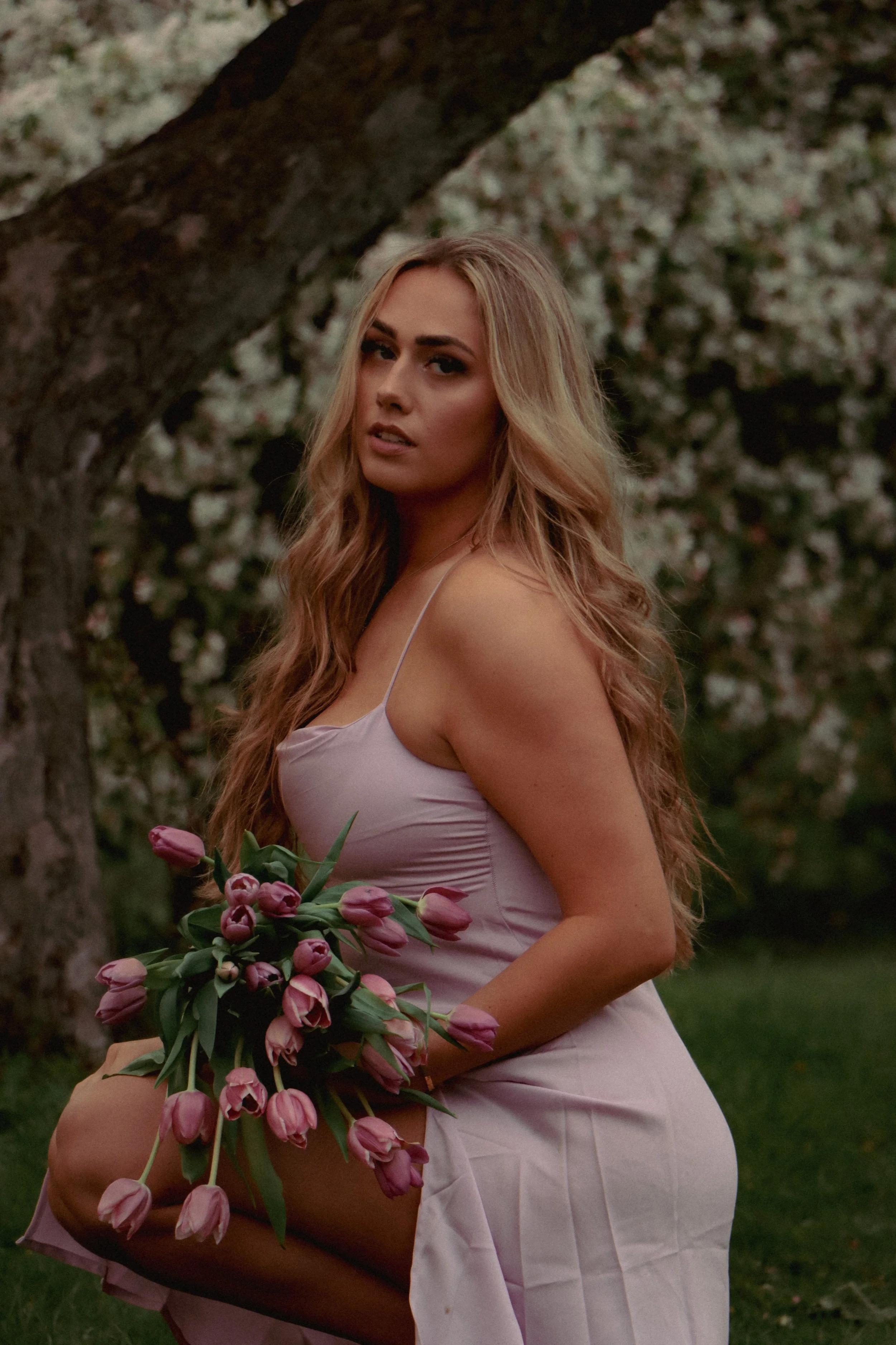 A woman with long, wavy blonde hair holding a bouquet of pink tulips, sitting outdoors near a flowering tree.