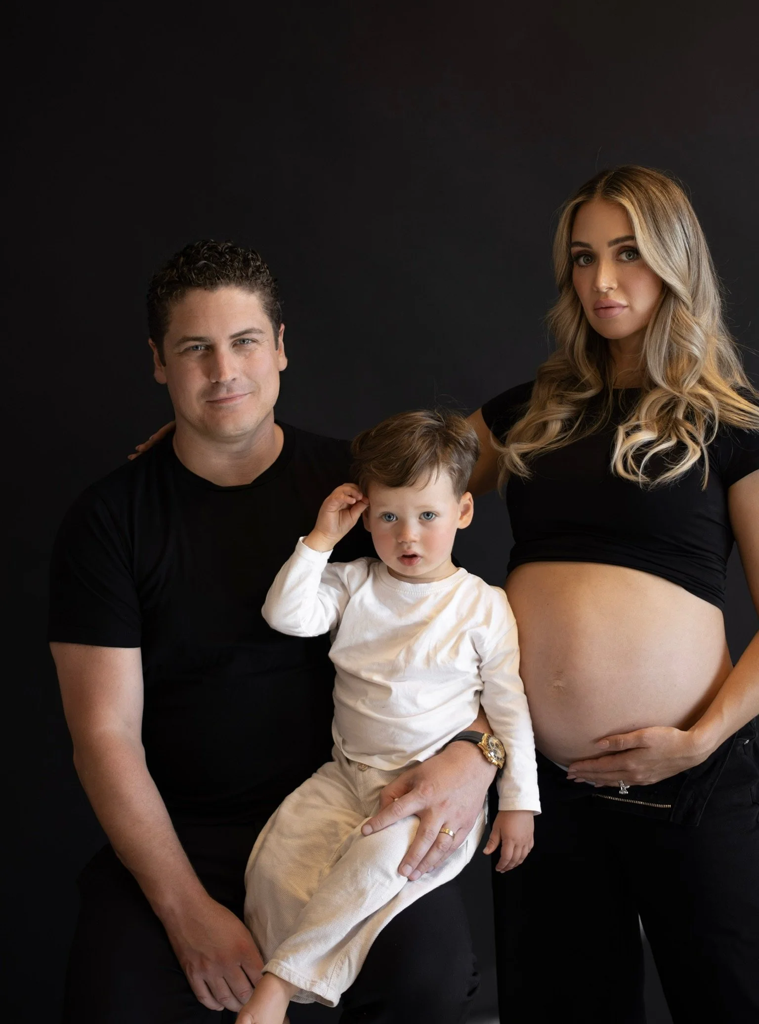 A family portrait of a man, woman, and young boy against a black background. The man has short curly hair and is wearing a black t-shirt. The woman has long, wavy blonde hair and is wearing a black crop top, showing her pregnant belly, with her hand 