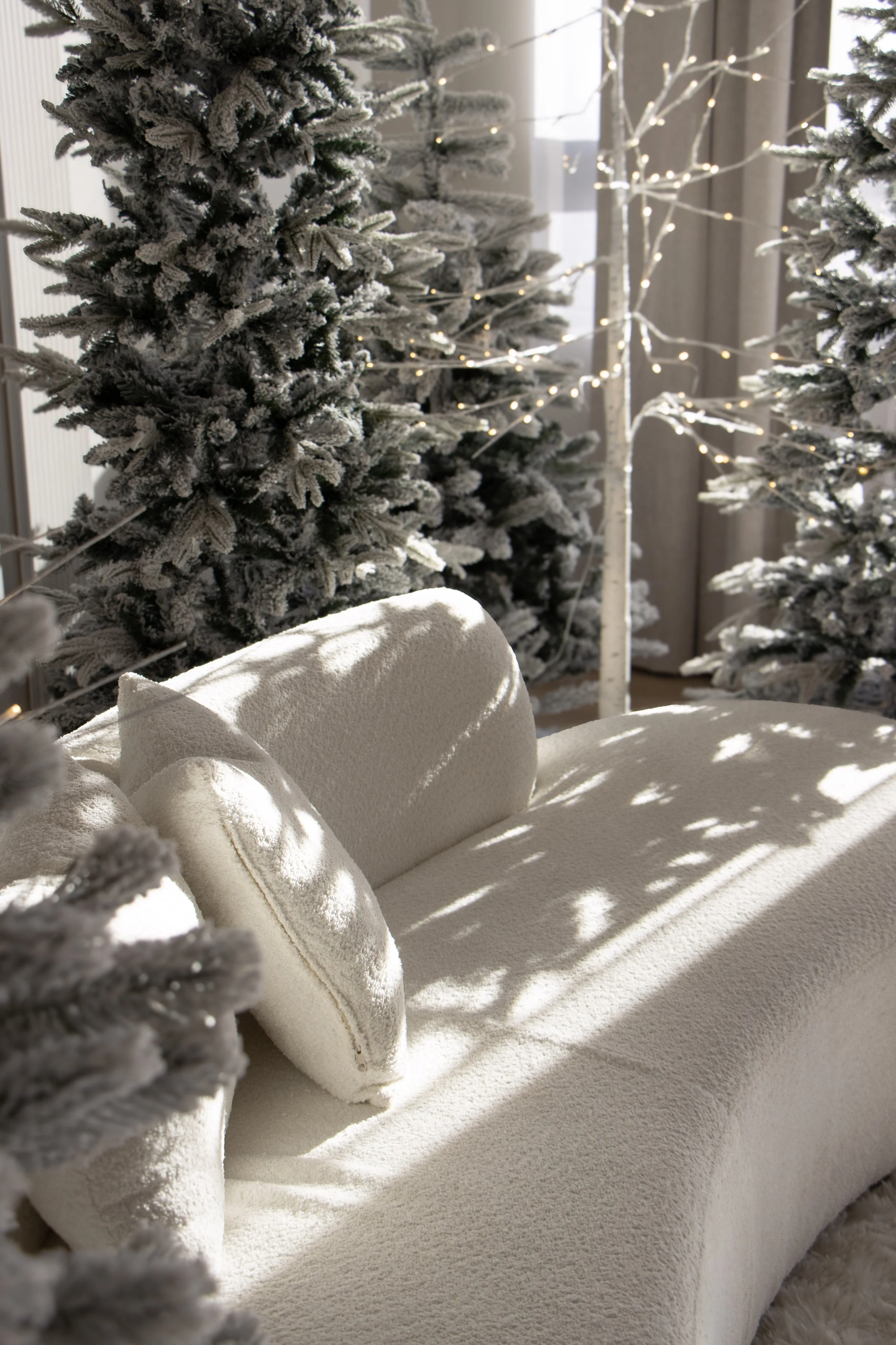 Snow-frosted holiday trees with fairy lights and a white textured sofa with cushions, illuminated by sunlight.
