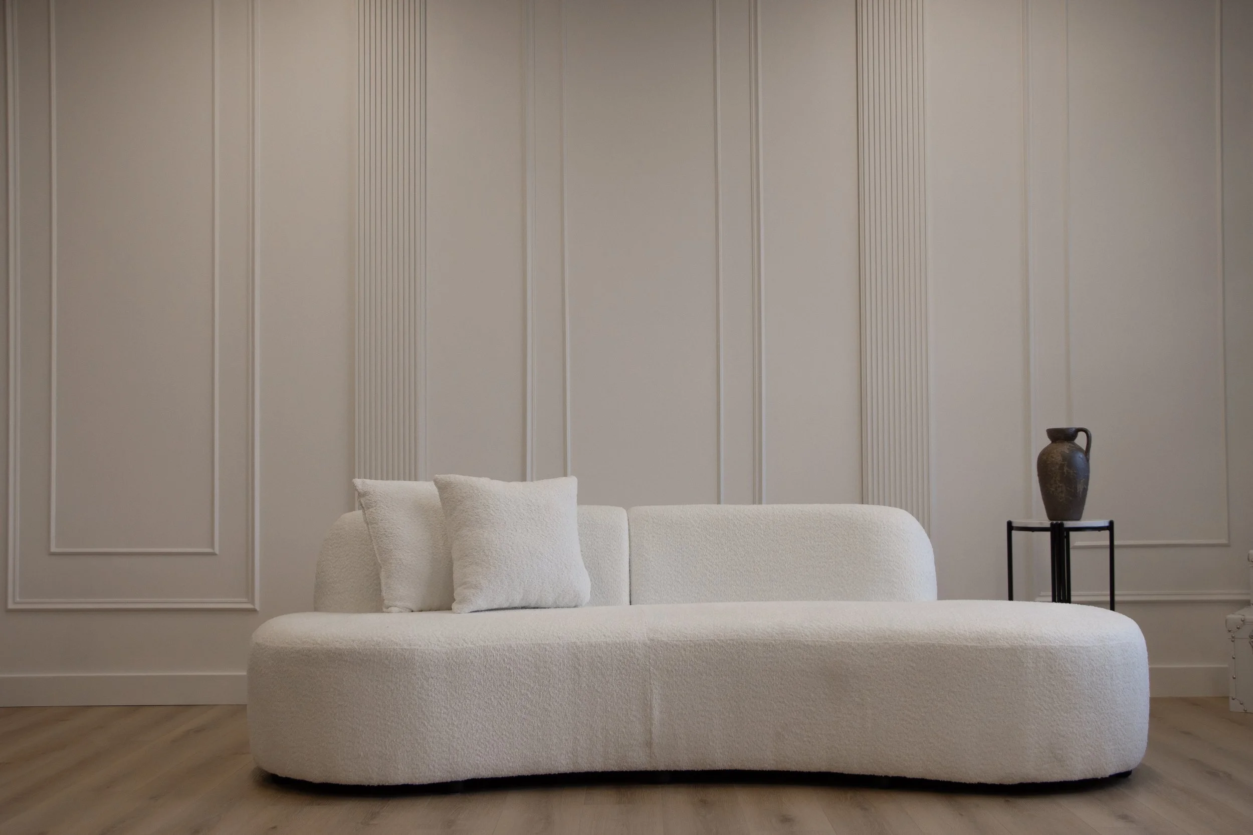 Minimalist living room with a white textured sofa, three white cushions, cream-colored wall paneling, and a small black side table with a dark ceramic vase.