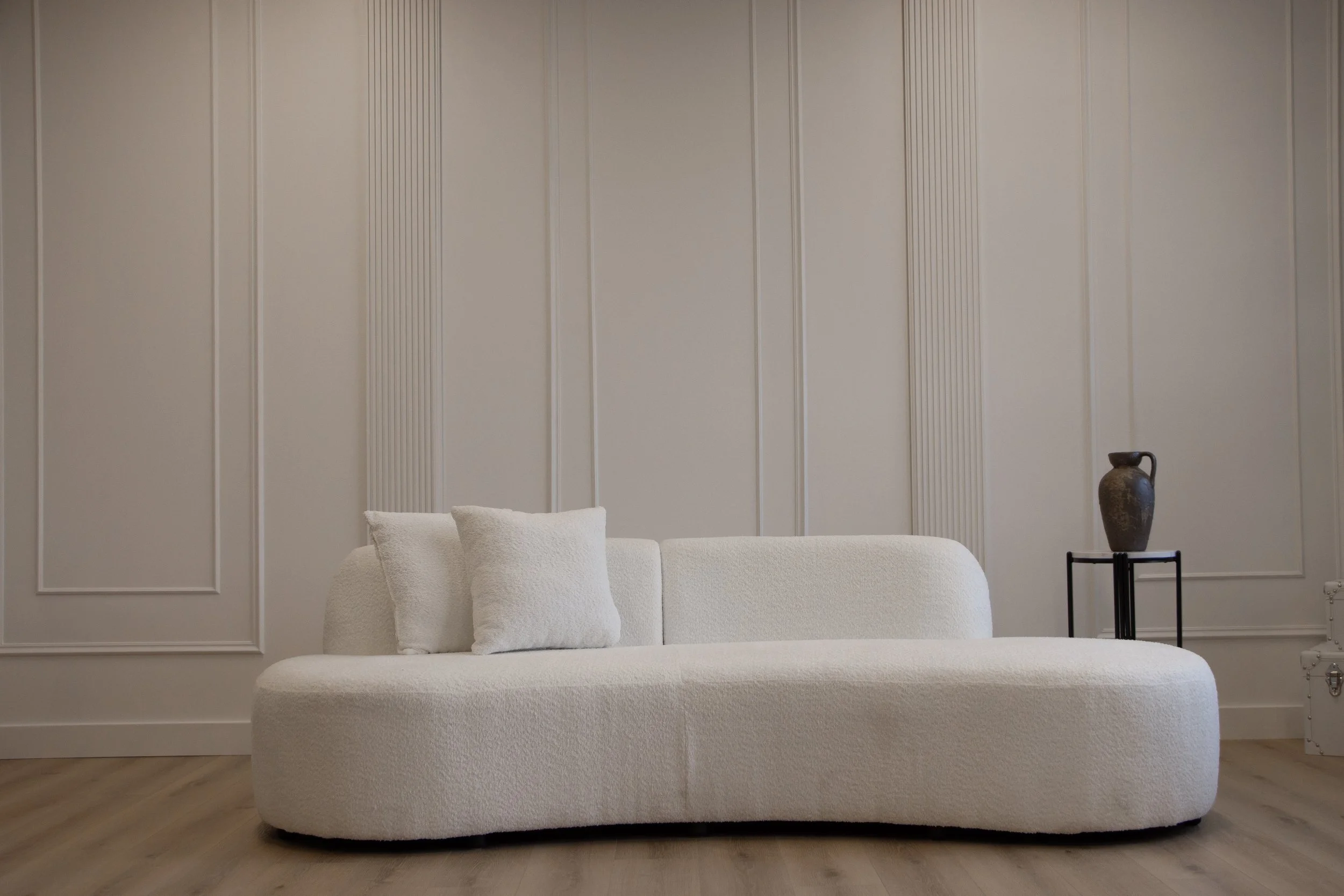 Minimalist living room with a white, textured sofa, two white cushions, a decorative vase on a black stand, and a metal trunk against a light paneled wall with wooden flooring.