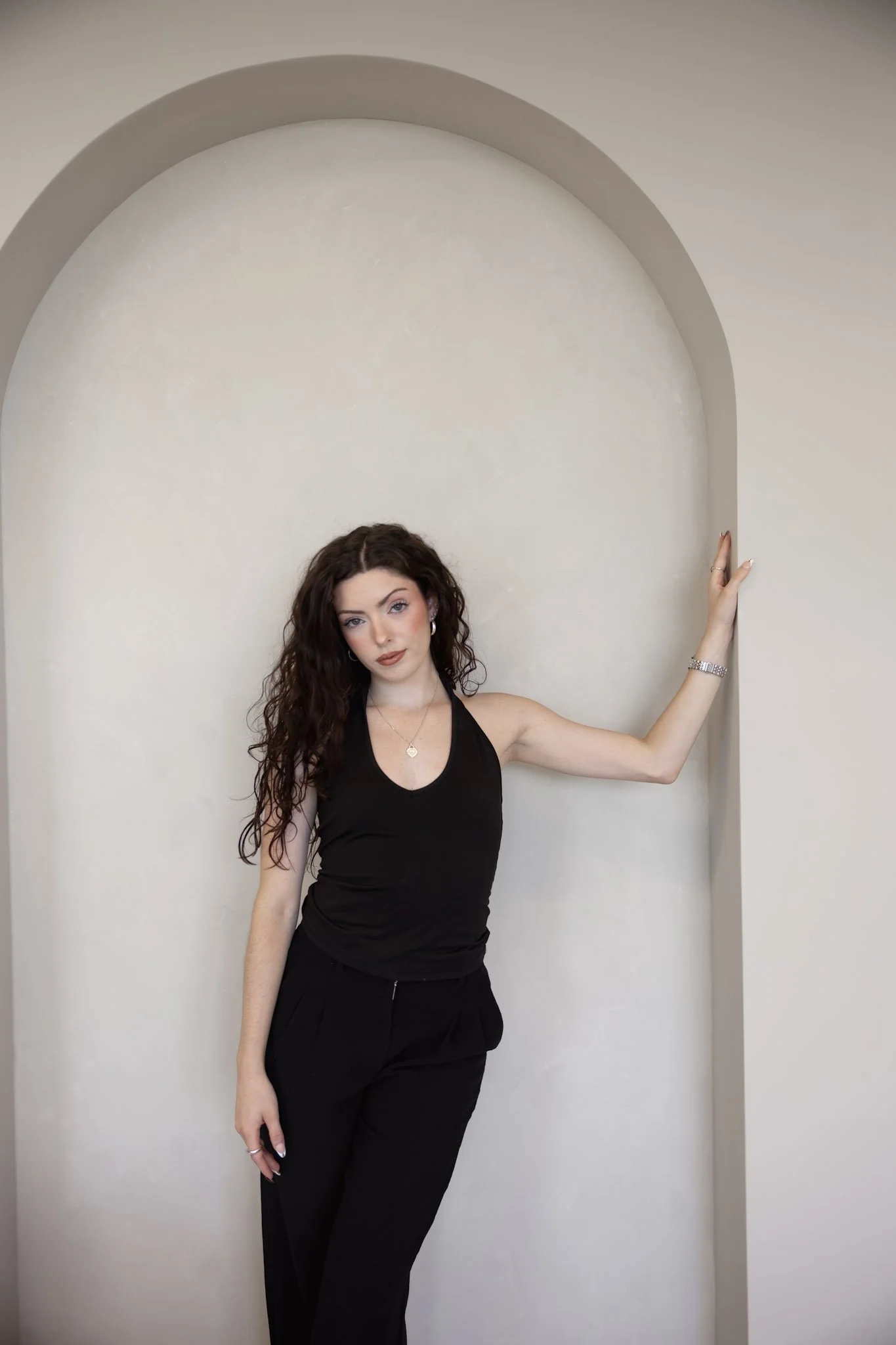 A woman with long, curly brown hair posing against a white wall with an arched alcove, wearing a black sleeveless top and black pants.