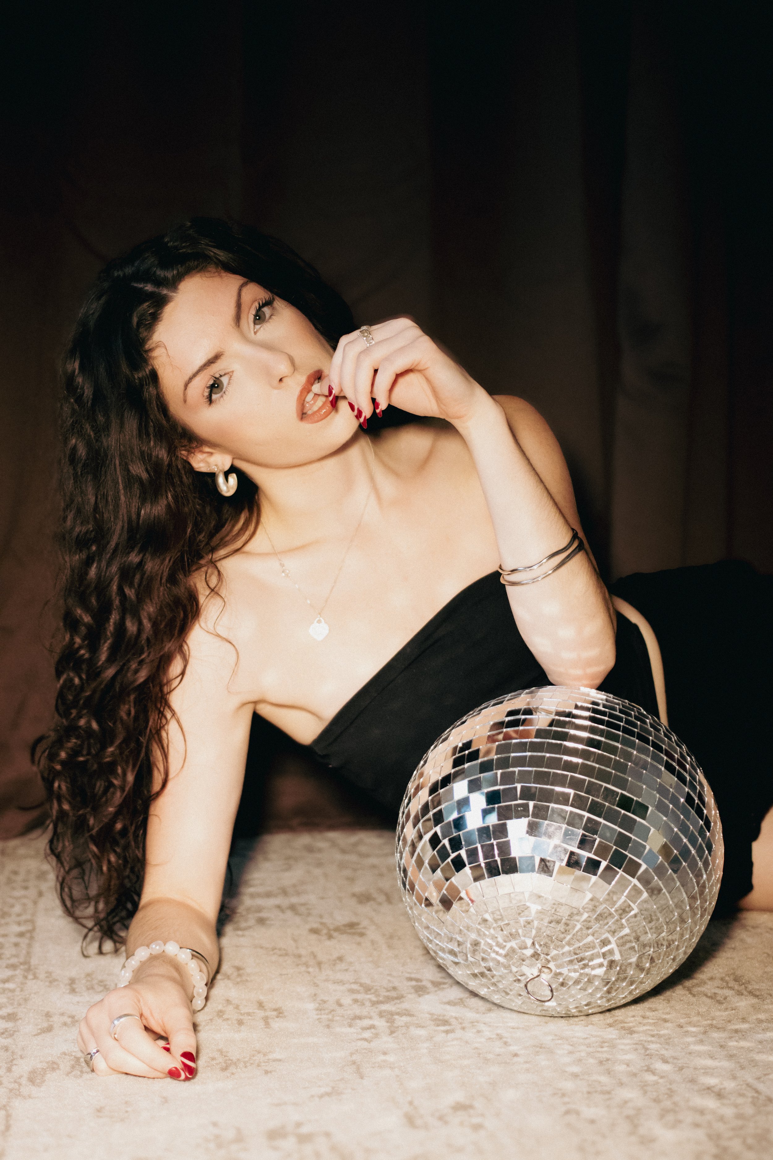 A woman with long, curly dark hair lying on a plush sofa, holding her finger near her lips, with a disco ball in front of her.