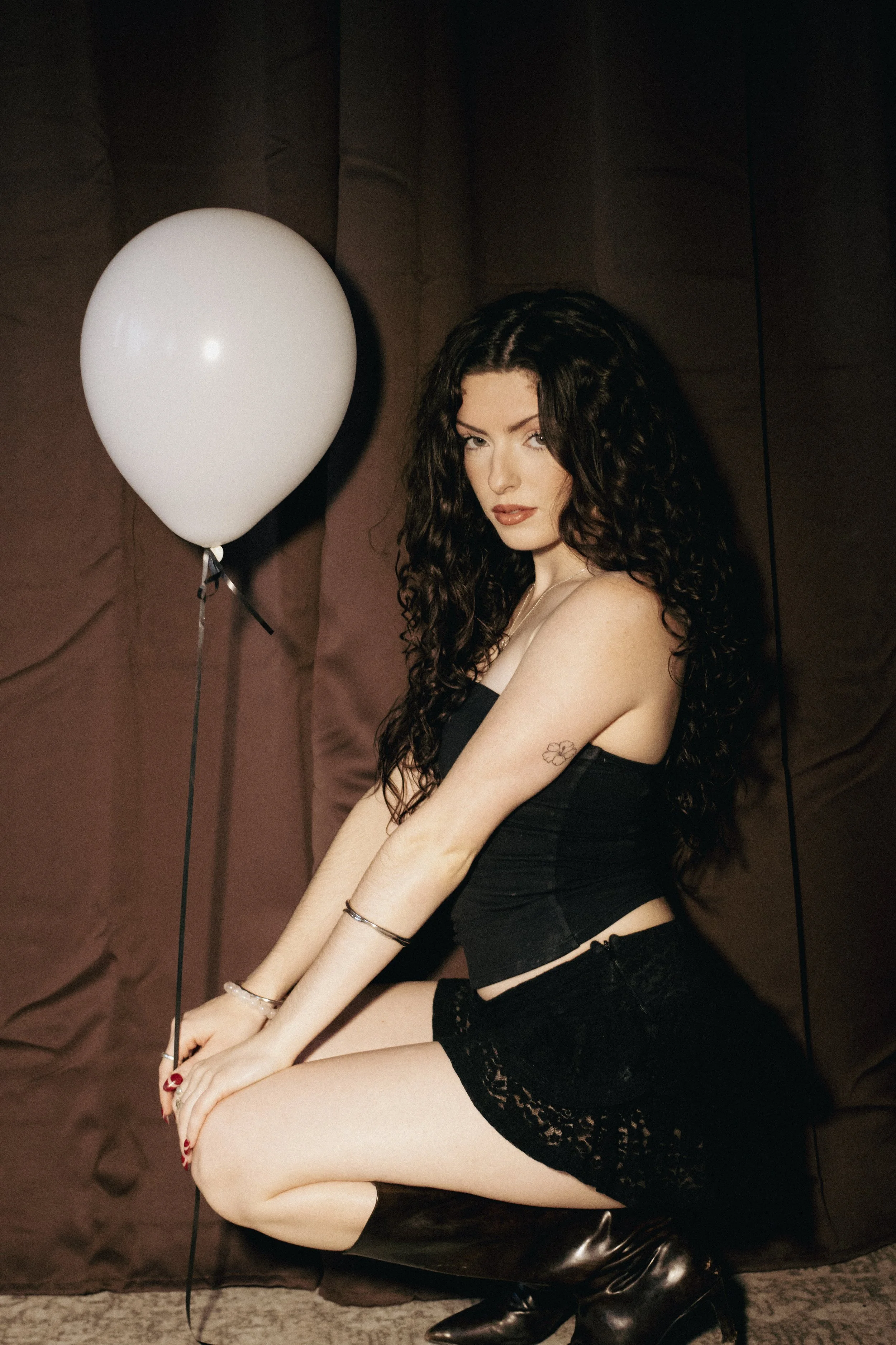 A woman with long curly dark hair sitting on a black stool, holding a white balloon, against a dark brown fabric backdrop.