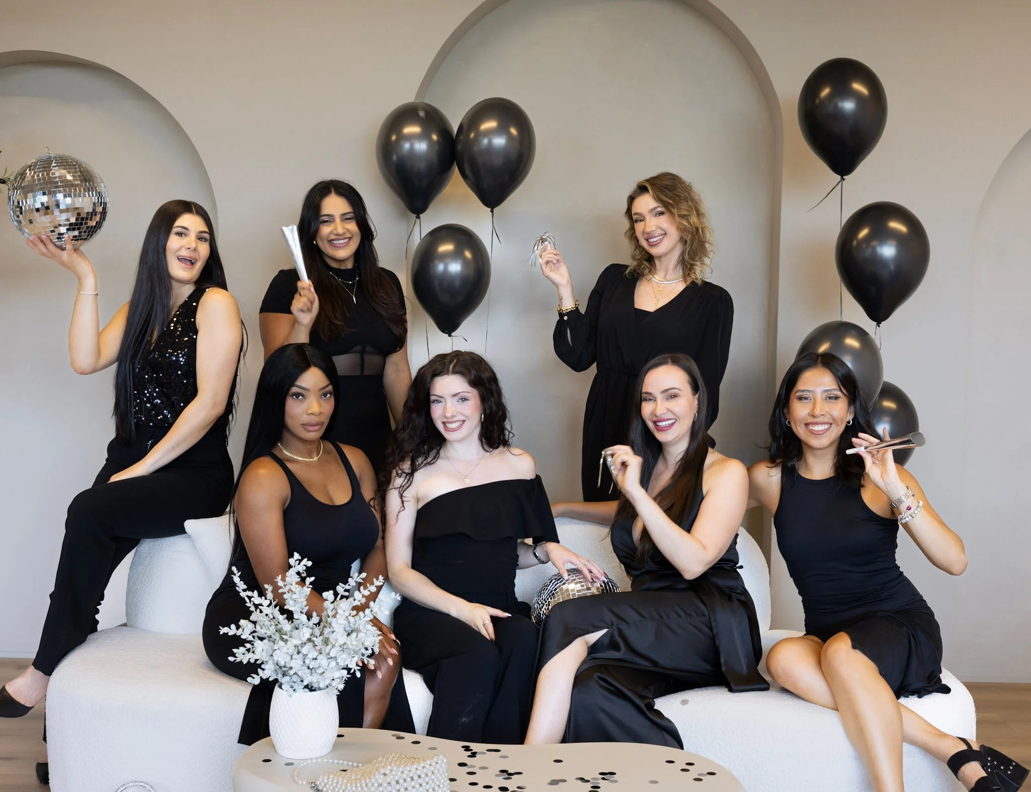 A group of eight women celebrating in black outfits with balloons and party accessories in a living room.