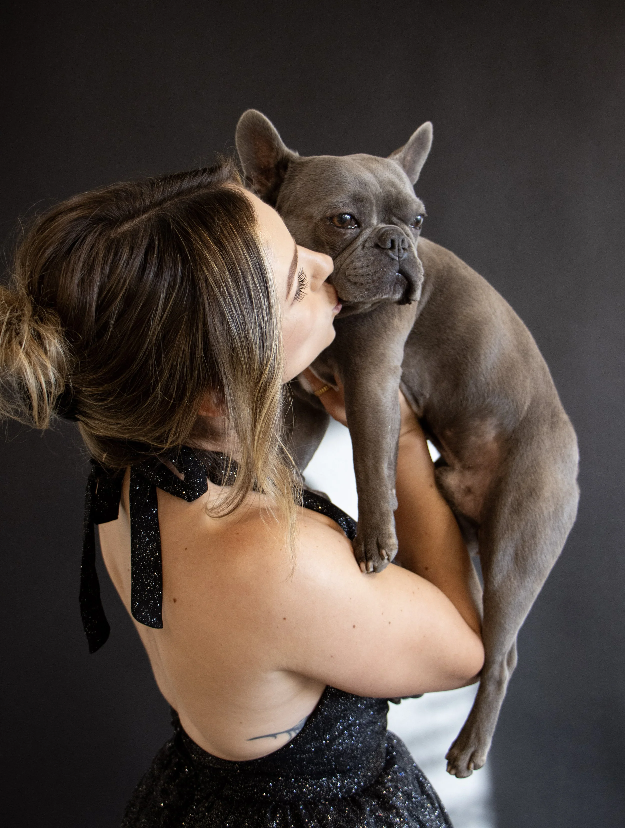 A woman holding a French Bulldog puppy close to her face, appearing to kiss or nuzzle it, against a dark background.