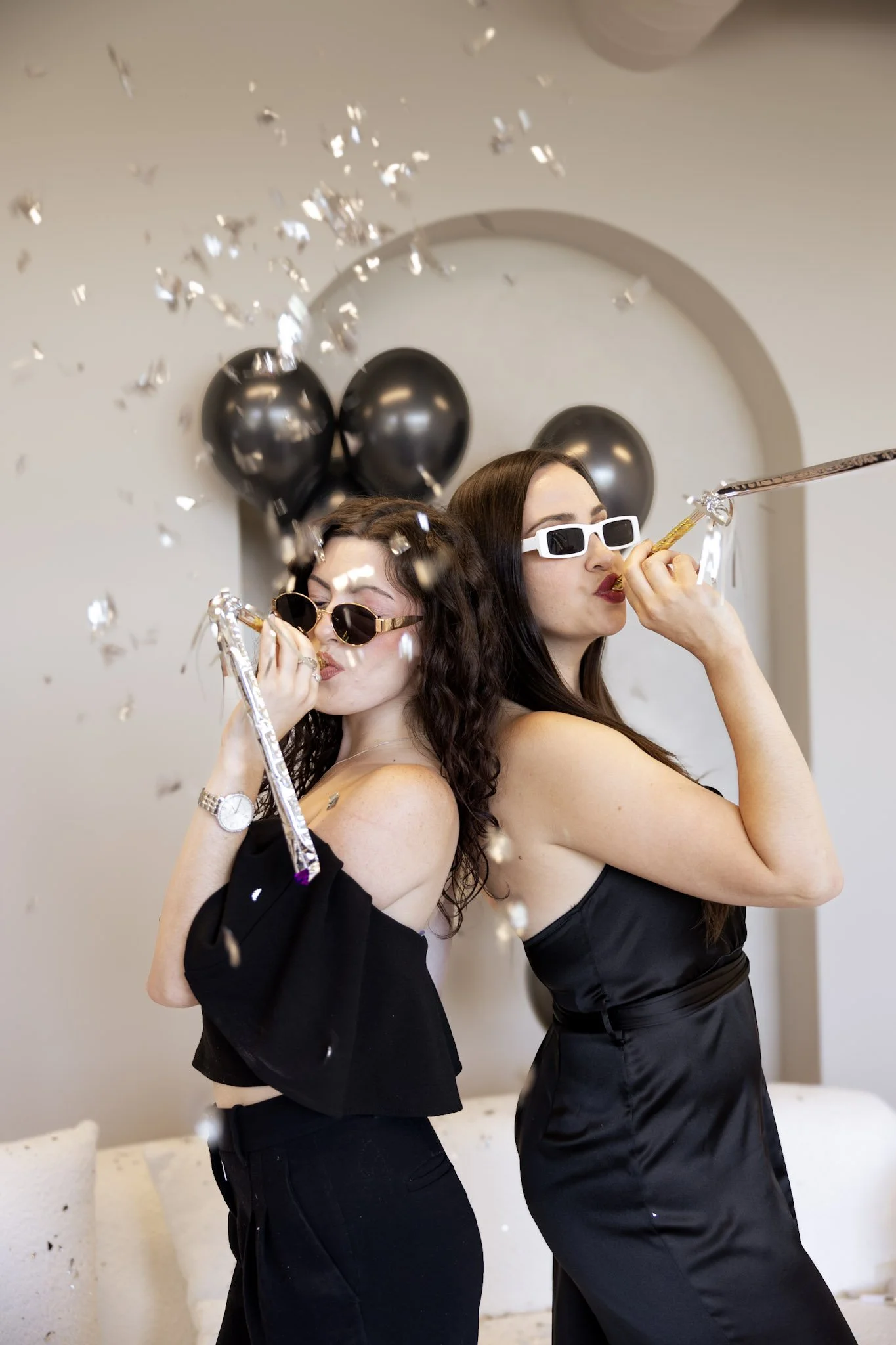 Two women celebrating with party blowers, black balloons, and confetti in a festive setting.