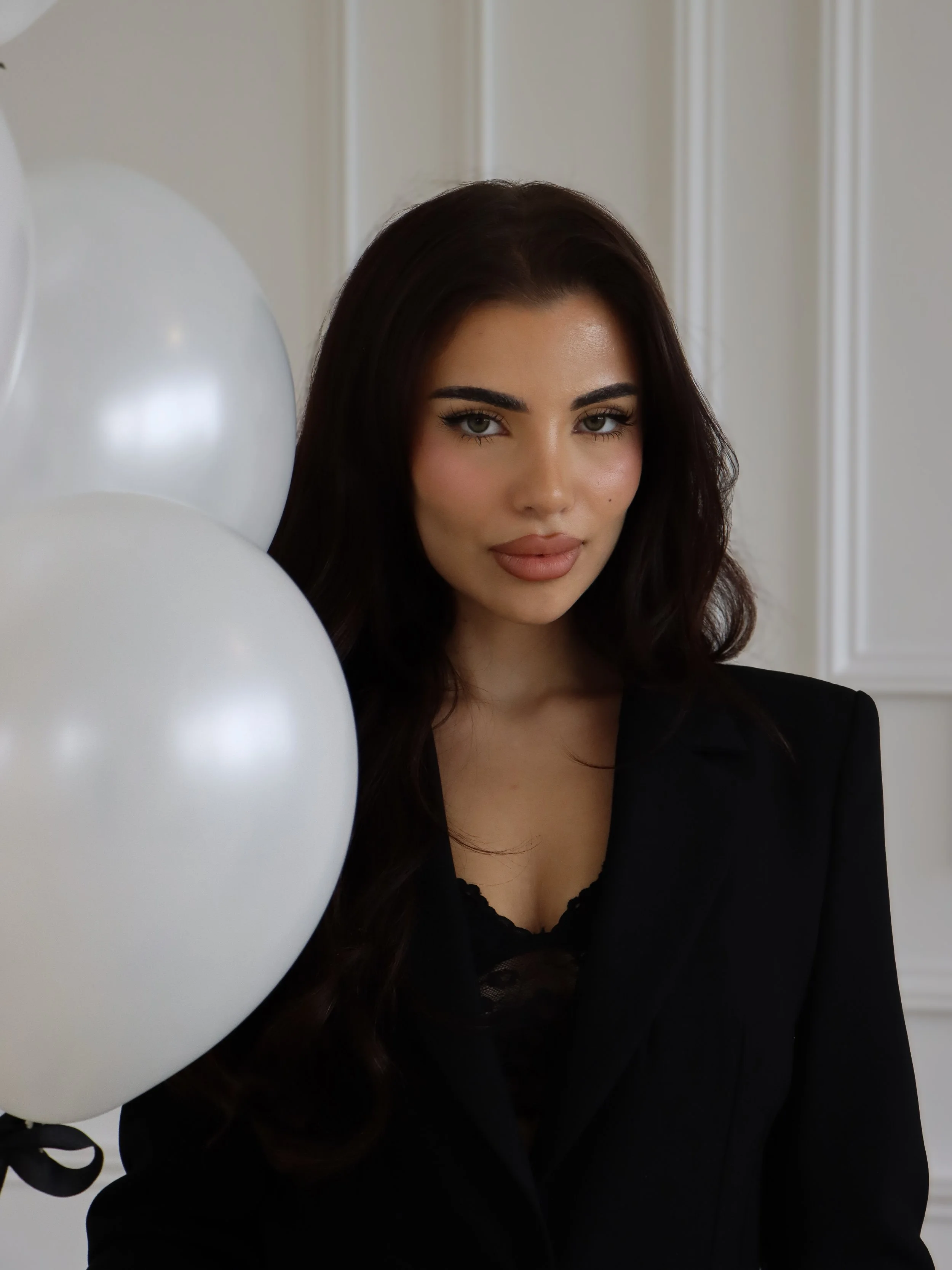 A woman with dark hair and makeup wearing a black blazer standing near a cluster of white balloons inside a room with white walls.