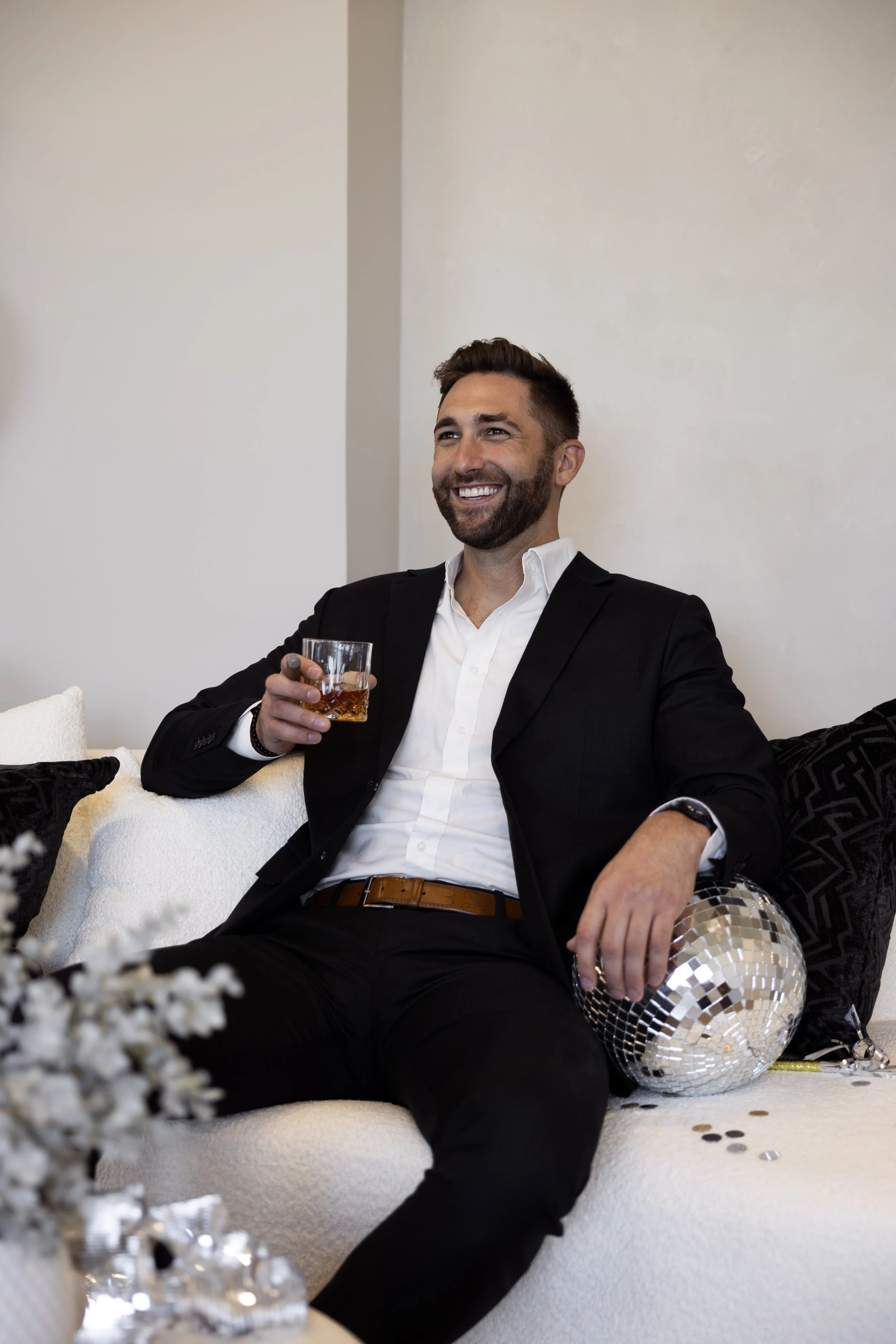 A man in a black suit and white shirt sitting on a white sofa, holding a glass of whiskey, smiling, surrounded by black pillows, a disco ball on his lap, and scattered confetti.