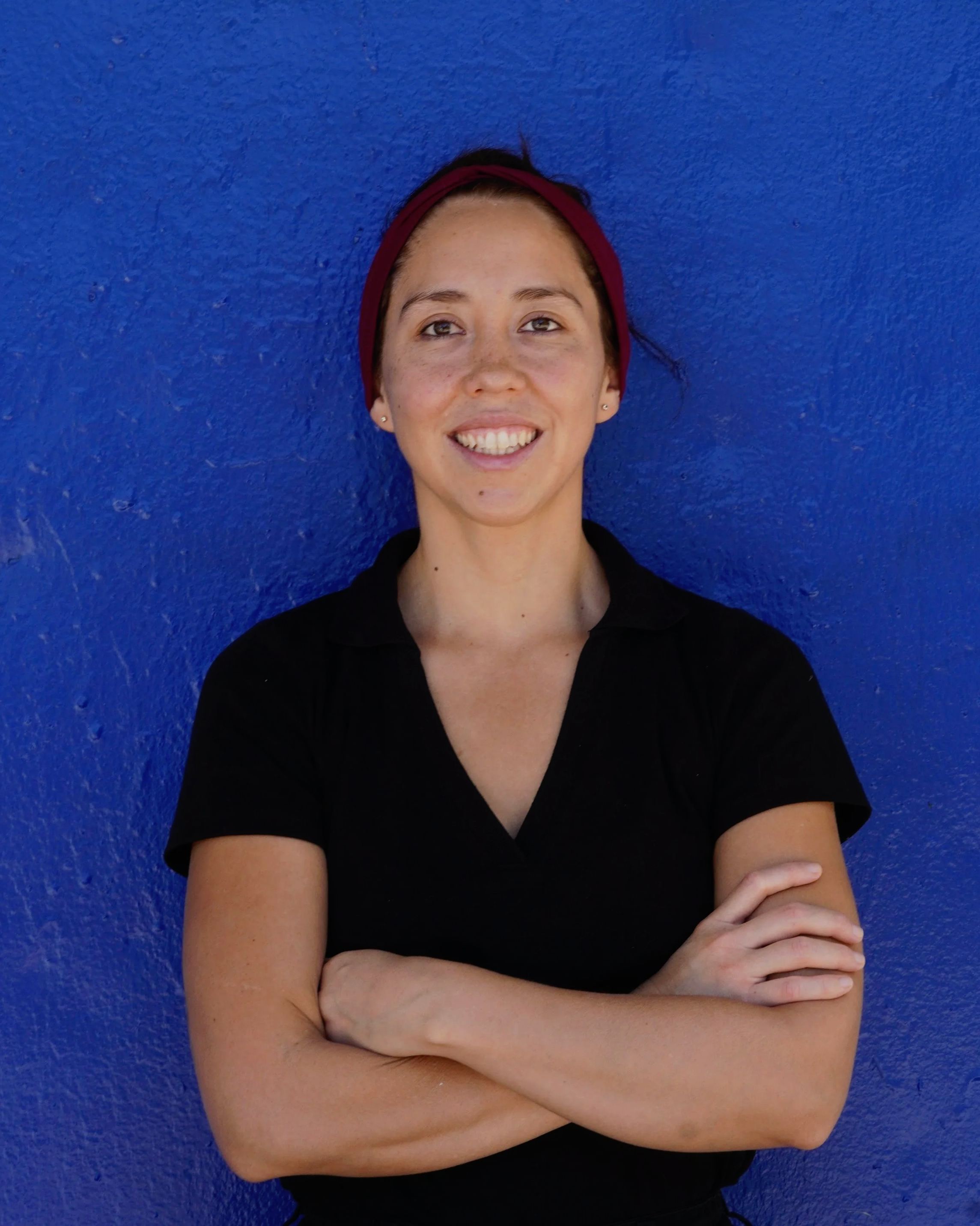 A young woman with short dark hair wearing a maroon headband, smiling, and crossing her arms, standing against a vibrant blue wall.