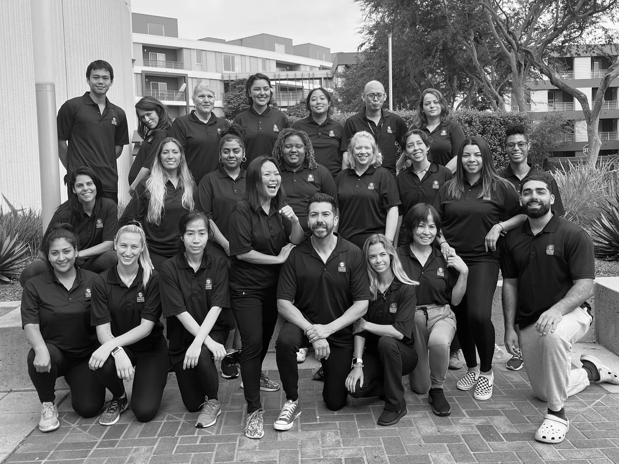 Group of diverse young people standing outdoors in front of modern apartment buildings, smiling and posing for a photo, some kneeling and some standing, with trees and bushes in the background.