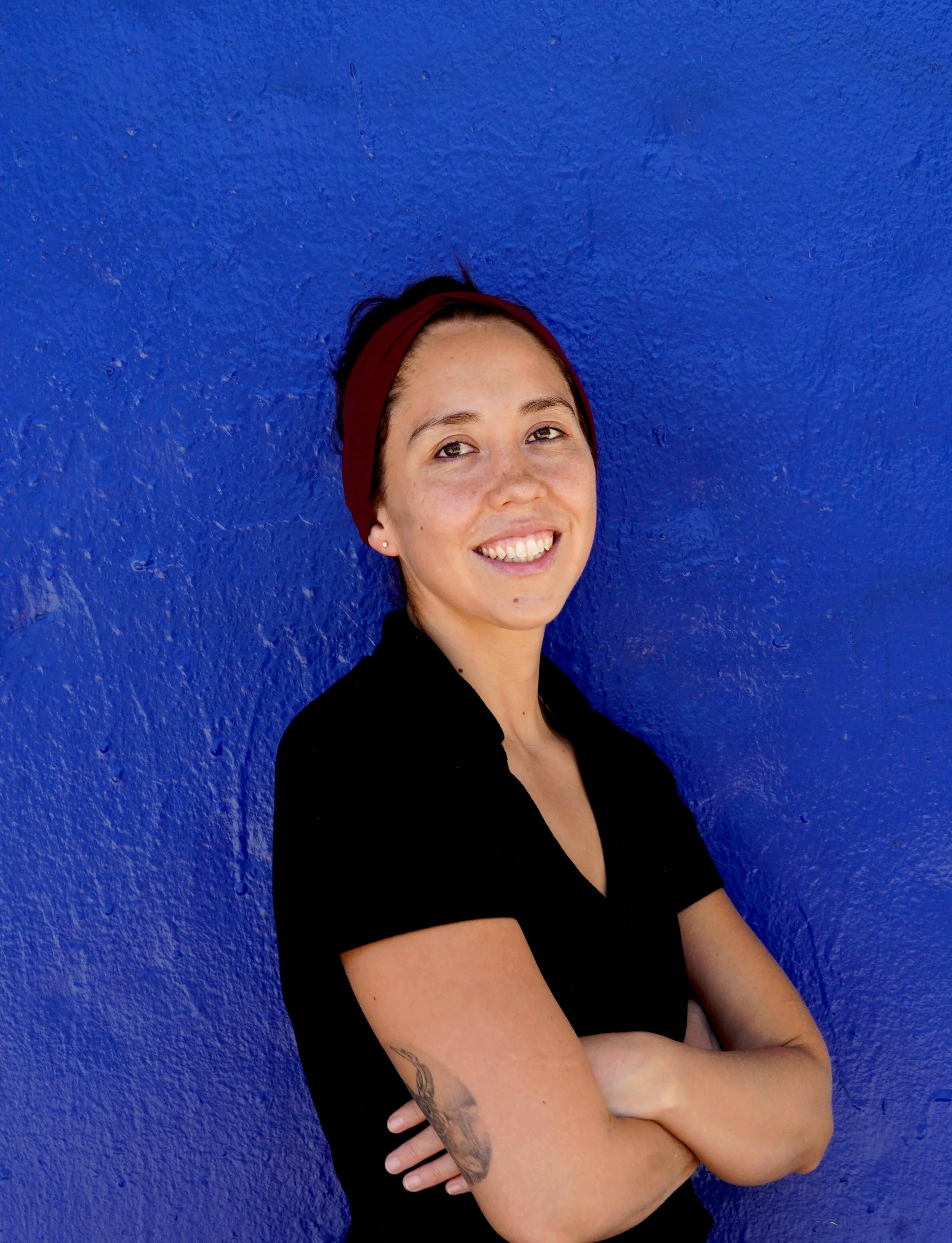 A woman with a red headband, short brown hair, smiling with a blue wall in the background, wearing a black shirt, with tattoos on her arms.