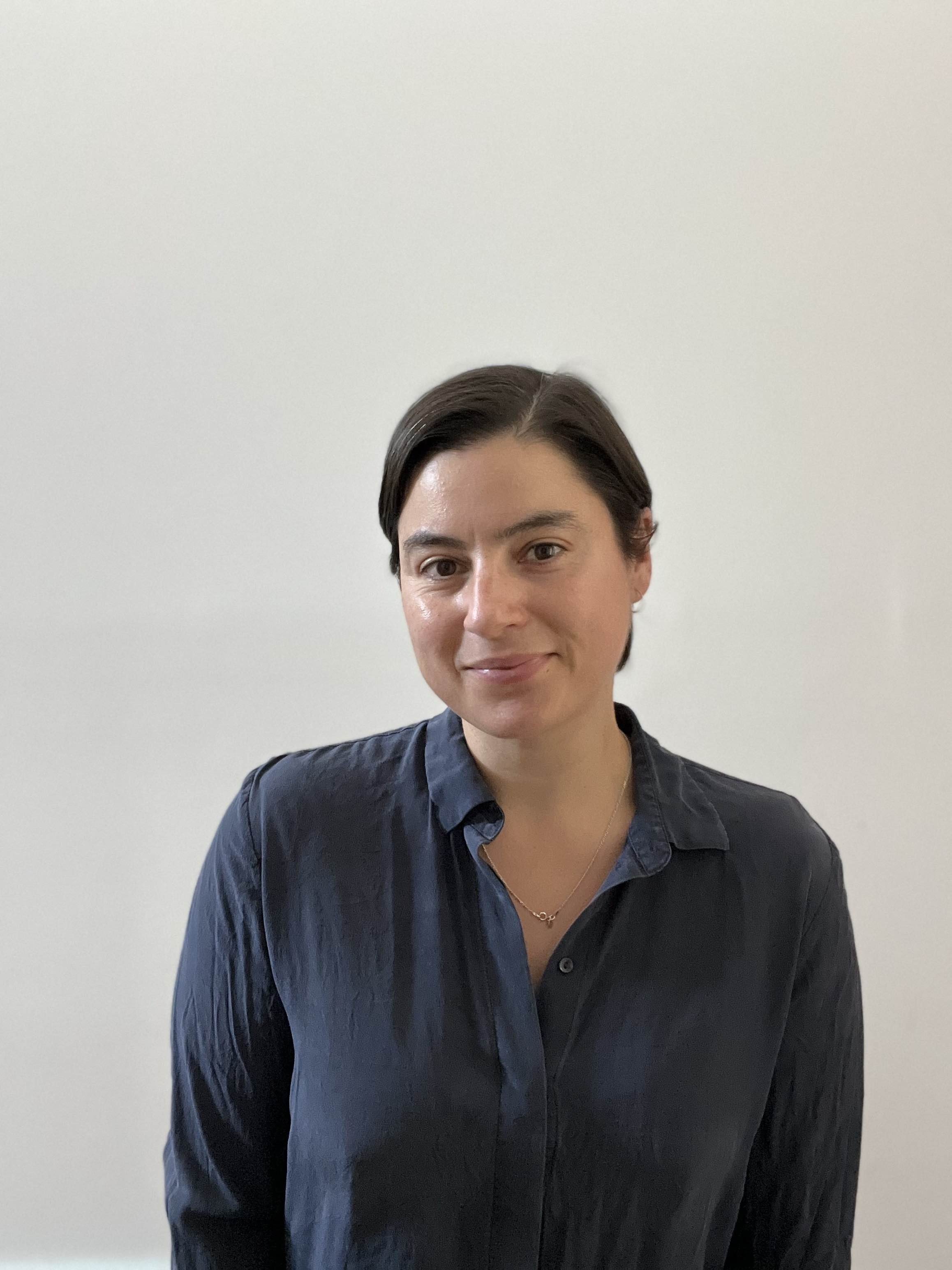 A woman with short dark hair wearing a dark blue button-up shirt standing against a plain white wall.