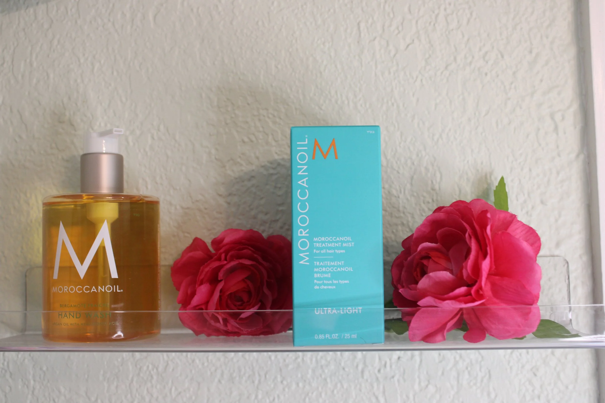 MOROCCANOIL hand wash bottle and treatment mist on a glass shelf with pink roses in a clear vase against a light-colored textured wall.