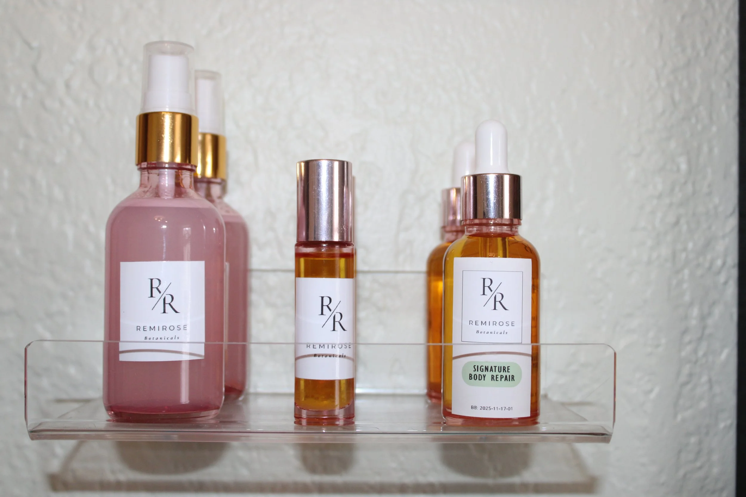 A clear wall-mounted shelf holding four skincare bottles with labels from Remirose Botanicals, including a pink spray bottle, a small amber roller bottle, another amber bottle with a dropper labeled 'Signature Body Repair,' and another pink bottle with a pump.
