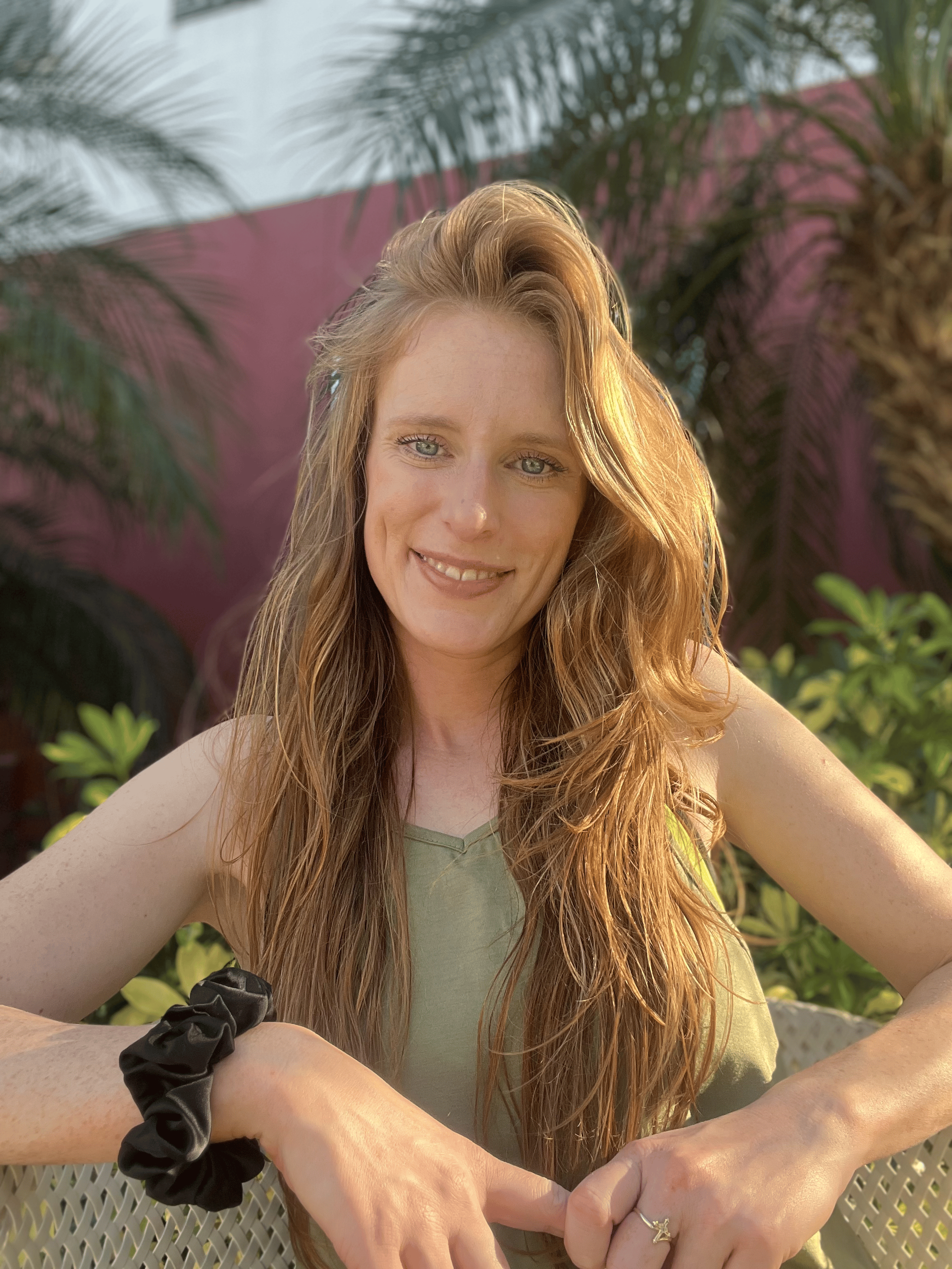 A woman with long red hair and blue eyes smiling outdoors, sitting at a table with green plants and a pink wall in the background.