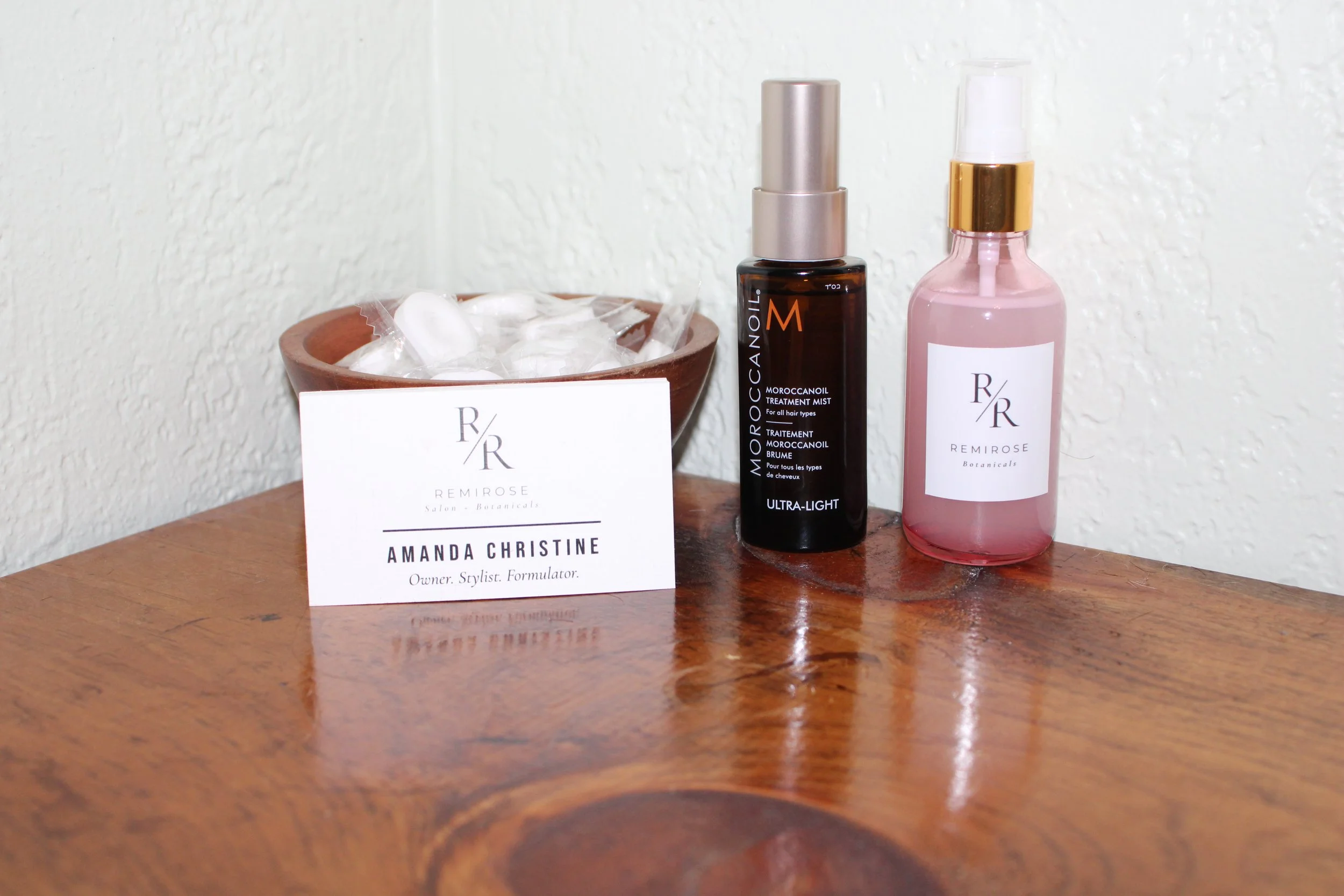 Business card reading 'Amanda Christine, Owner, Stylist, Formulator' on a wooden table next to a small bowl with cotton swabs, a bottle of Moroccanoil Treatment Mist, and a bottle of Remi Rose Botanicals facial mist.