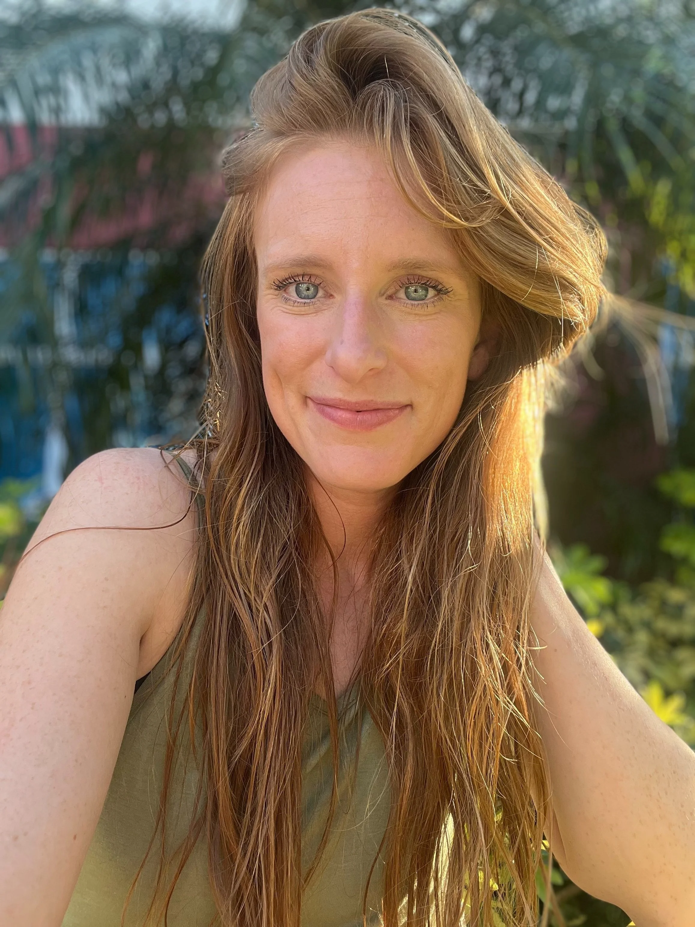 A woman with long, wavy red hair and blue eyes taking a selfie outdoors with lush greenery in the background.