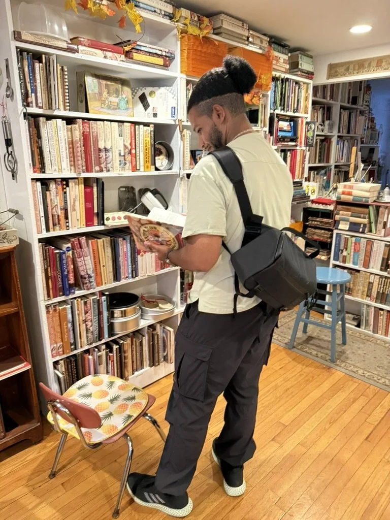 Man with a backpack reading a book in a bookstore or library, surrounded by shelves filled with books, with a small chair and a blue stool nearby.