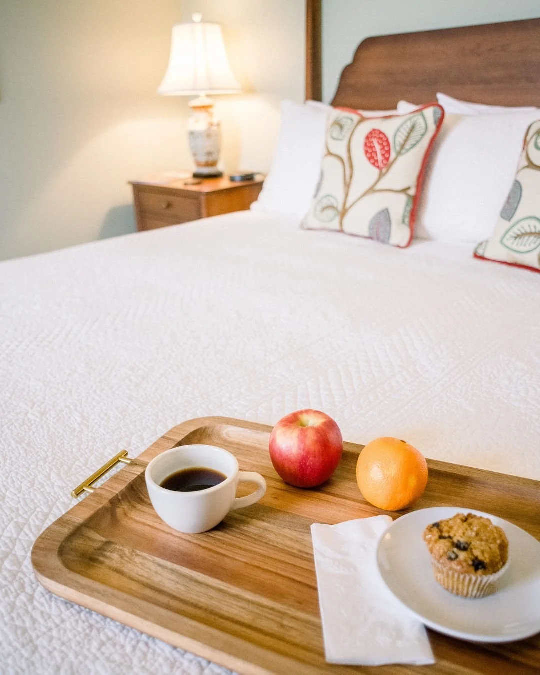 A tray with a cup of coffee, an apple, an orange, and a blueberry muffin on a bed with pillows in a cozy bedroom.