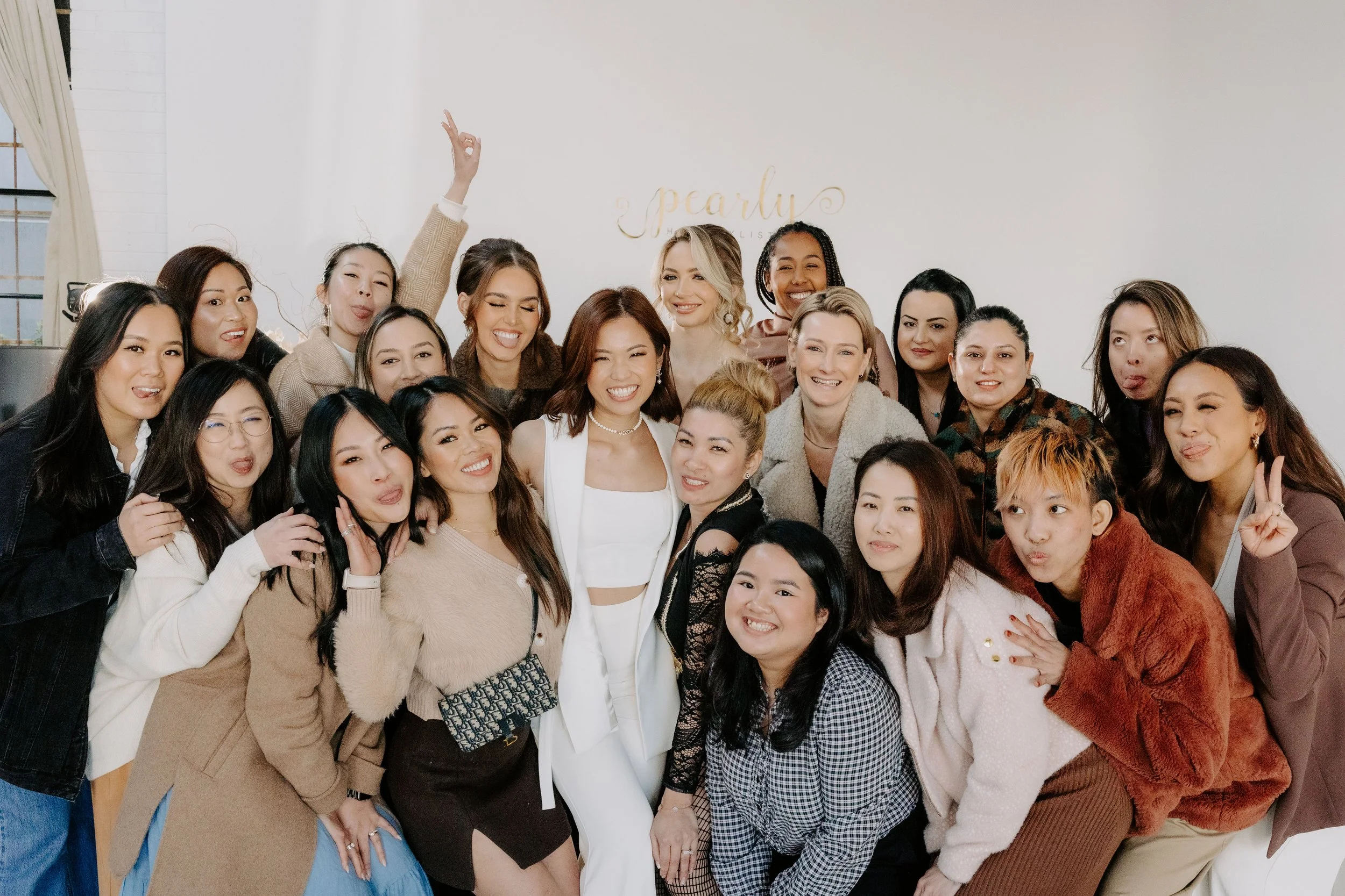 A diverse group of women posing together indoors, smiling and making playful gestures, in front of a white wall with the names Pearlby and a gold-colored script lettering.