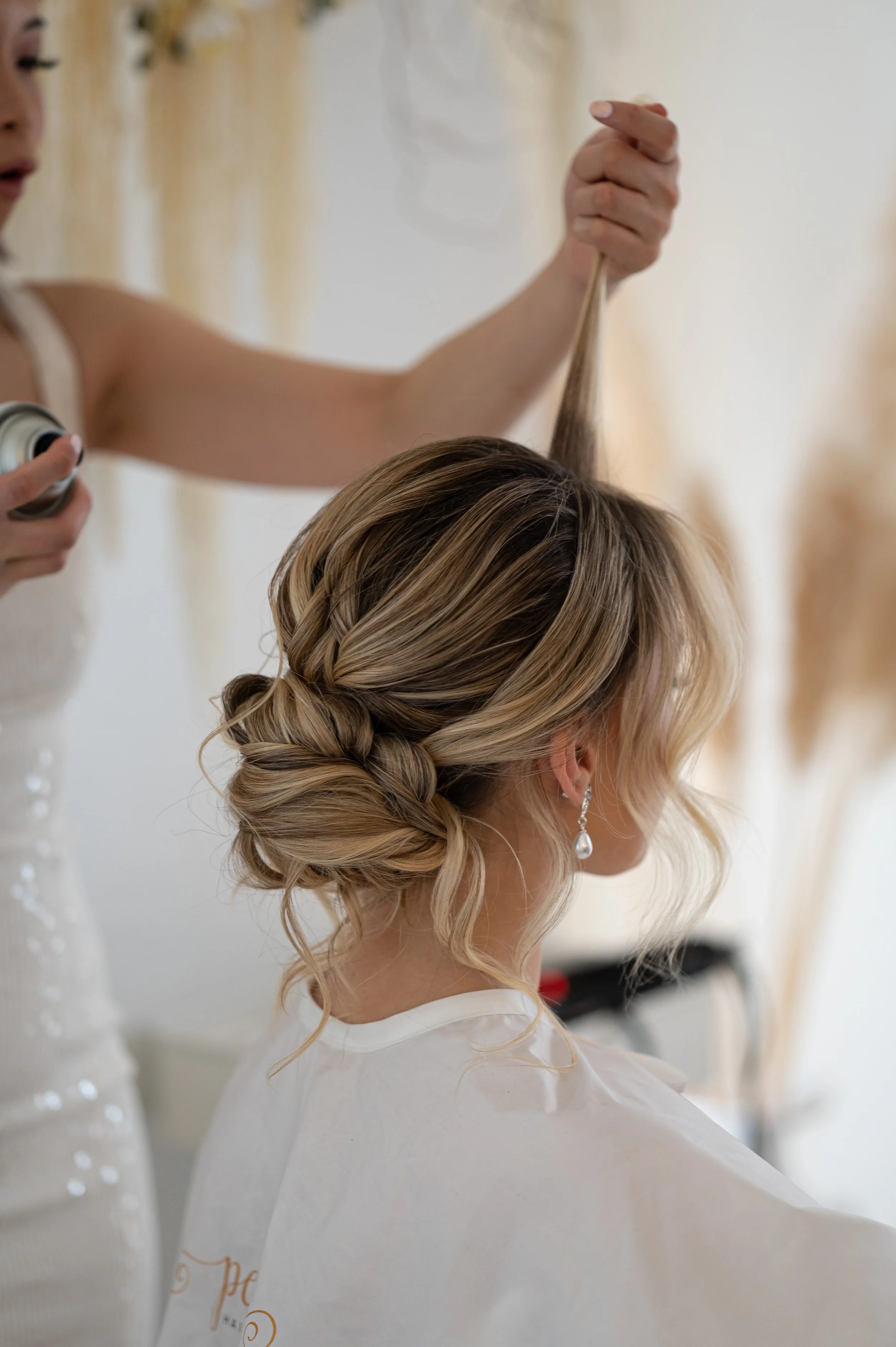 A woman with blonde hair styled in an elegant updo with braids is having her hairstyle done by a hairstylist who is spraying hair spray. The woman is wearing pearl earrings and a white cape.