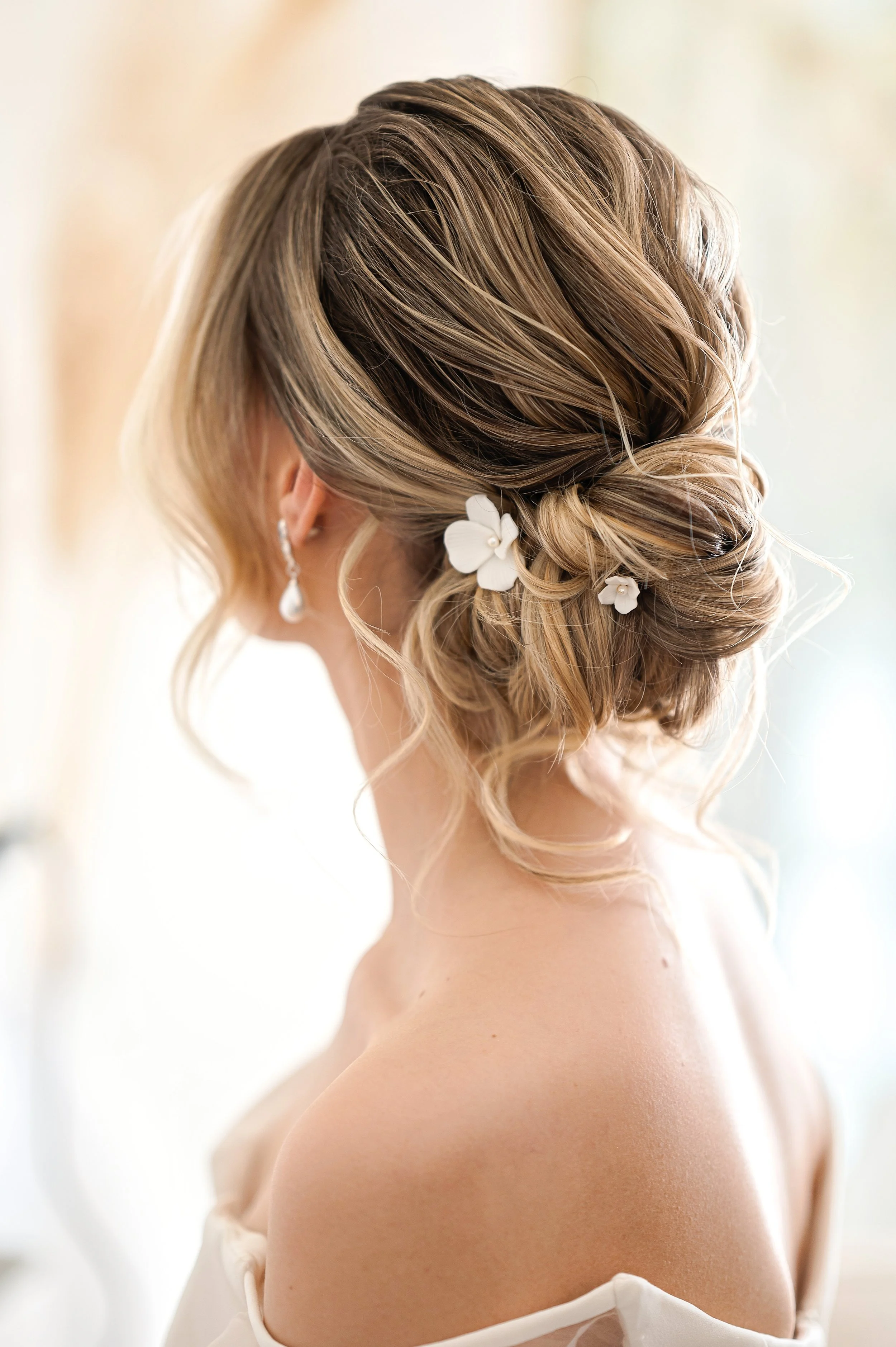 Close-up of a woman's styled updo hair with white flower hair accessories, highlighted with blonde tones, wearing pearl earrings and a white off-shoulder dress.