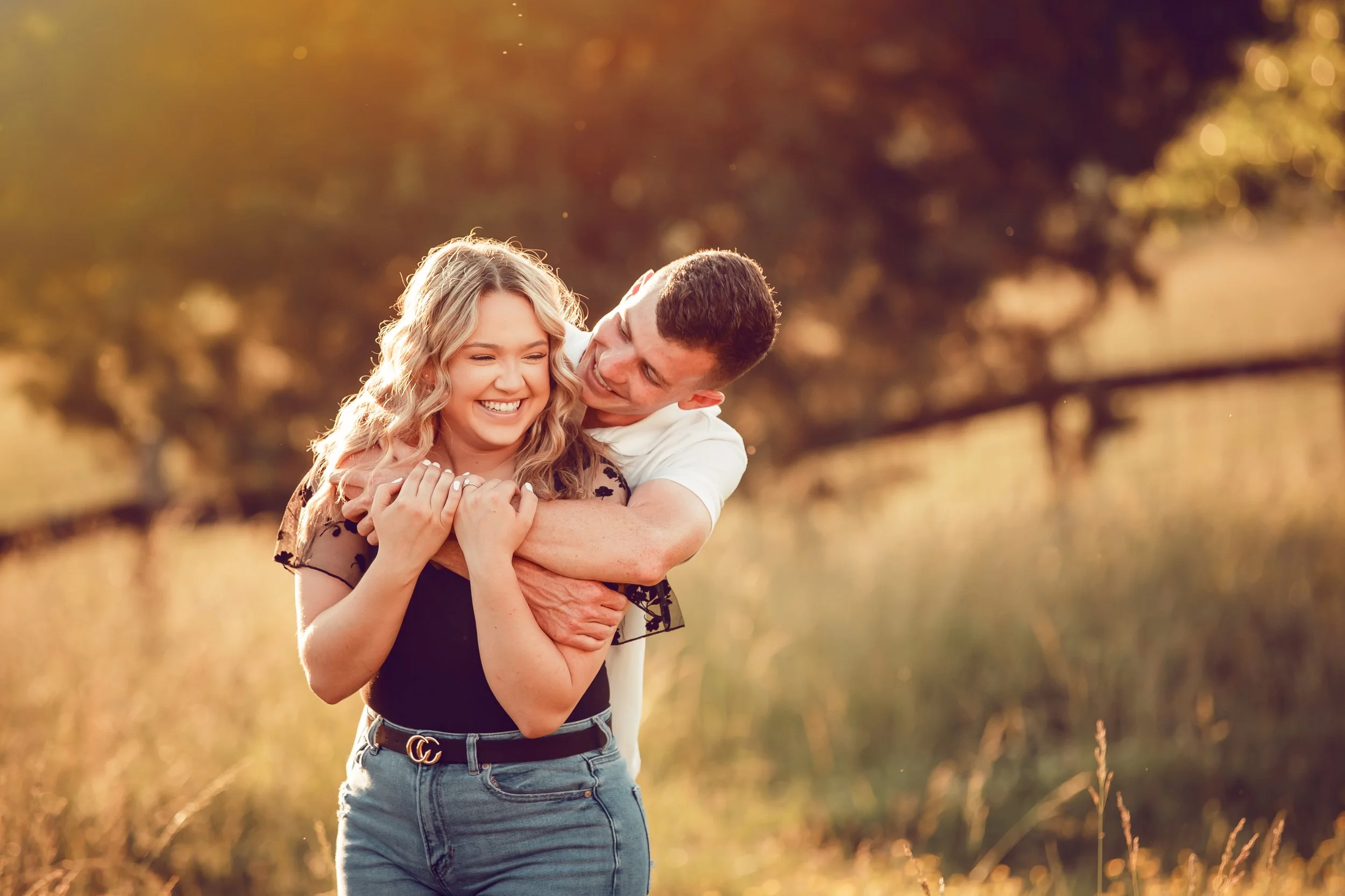 A young man hugging a young woman from behind outdoors during golden hour, both smiling and enjoying each other's company.