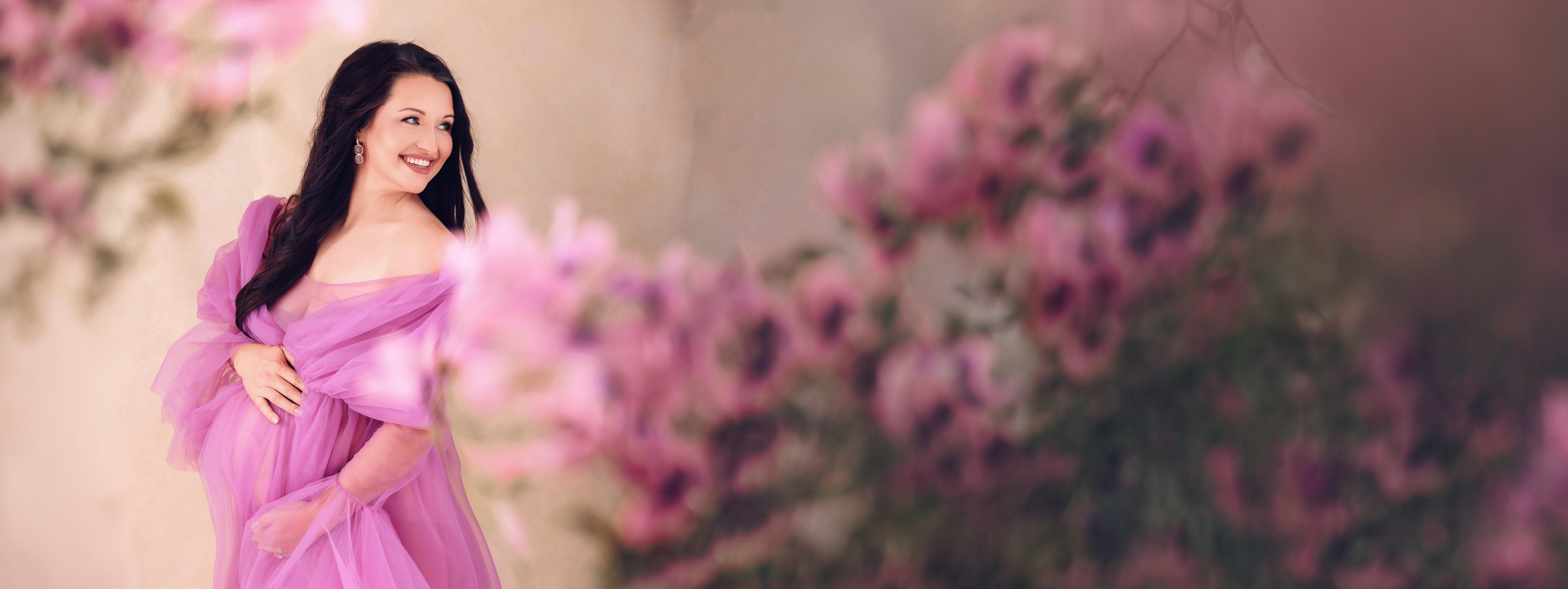 A woman wearing a flowing pink dress, smiling and looking to the side, with blurred pink flowers in the foreground.