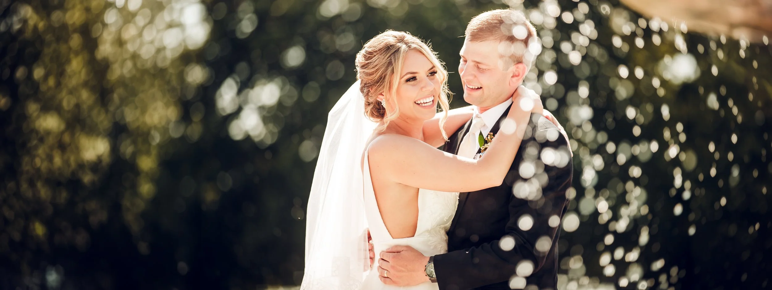 A bride and groom sharing a joyful embrace outdoors on their wedding day, with sunlight and bokeh background.
