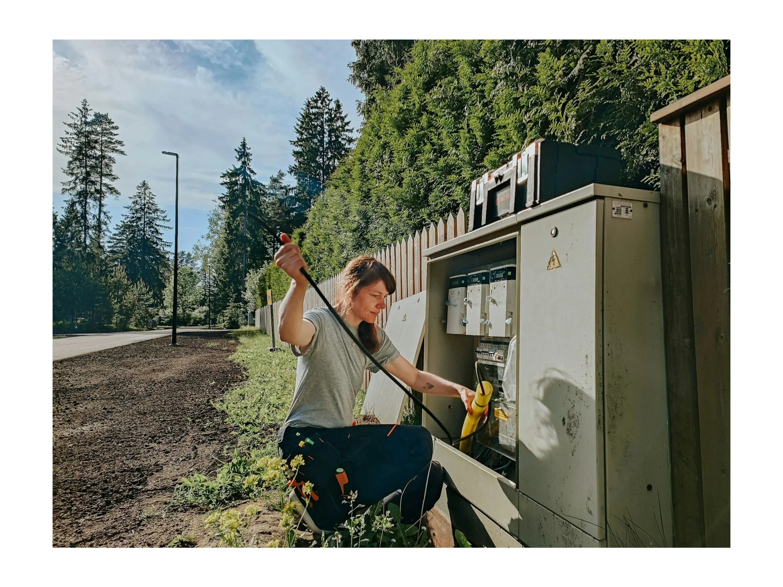 Frau repariert eine defekte Stromverteilerkasten im Freien, umgeben von Bäumen und einer Straße in einer ländlichen Gegend, bei sonnigem Wetter.