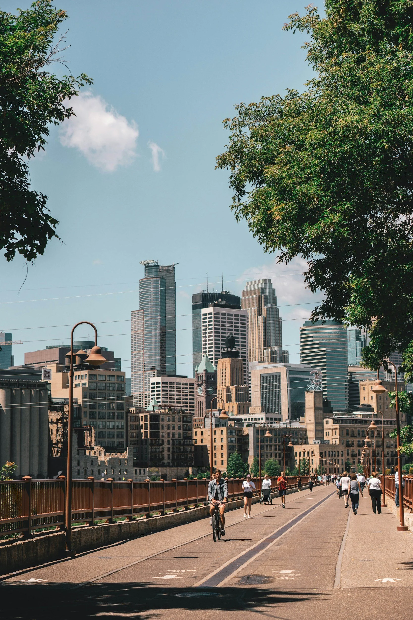 People walking and cycling on a city bridge with tall skyscrapers in the background, trees on the sides, and a clear blue sky.
