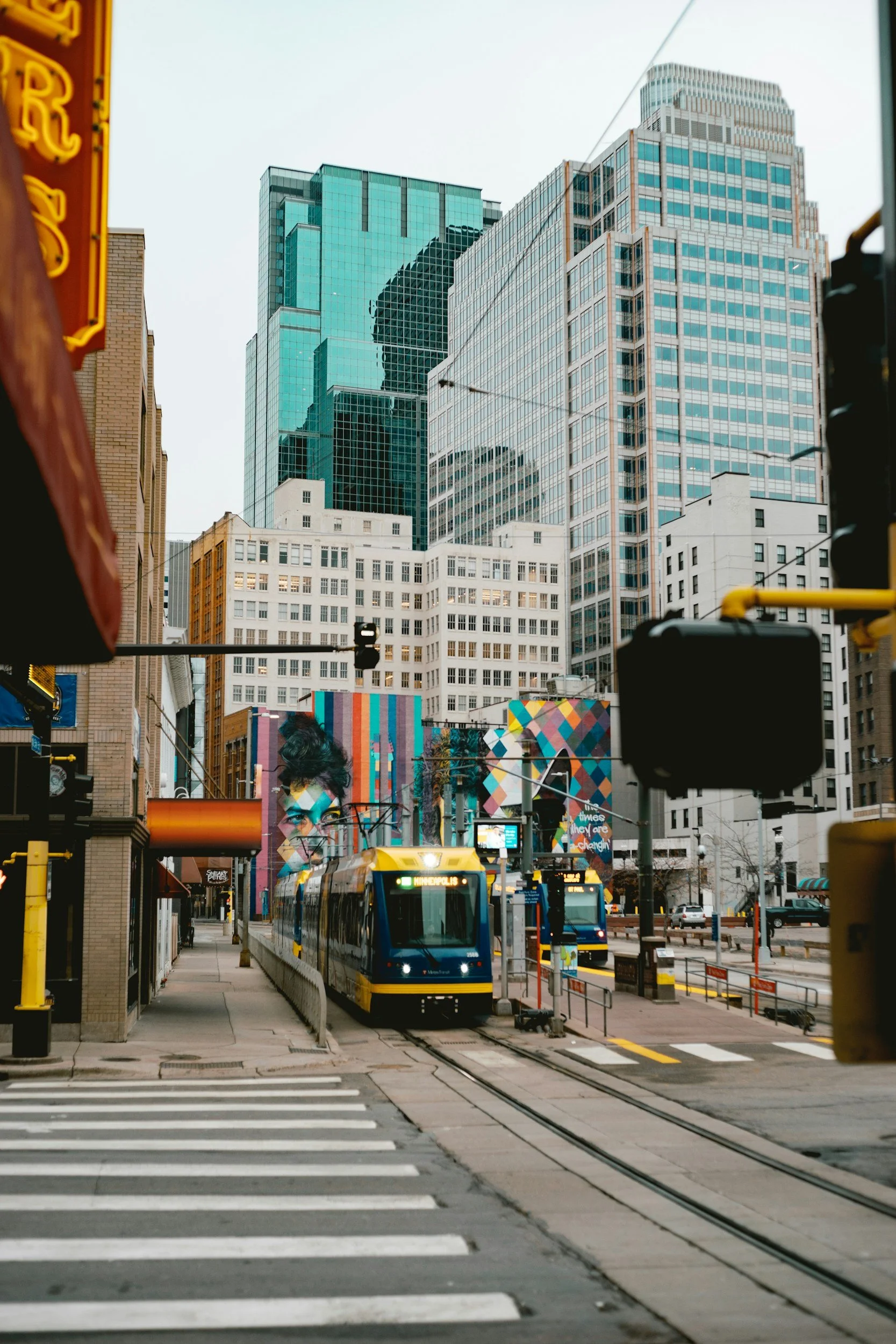 View of city street with modern high-rise buildings, a light rail train on tracks, and streetcars, with a crosswalk in the foreground.