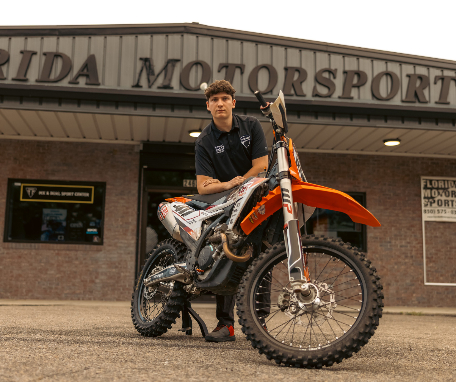 Young man standing behind an orange and white dirt bike in front of a building with a sign that reads 'Florida Motorsport'.