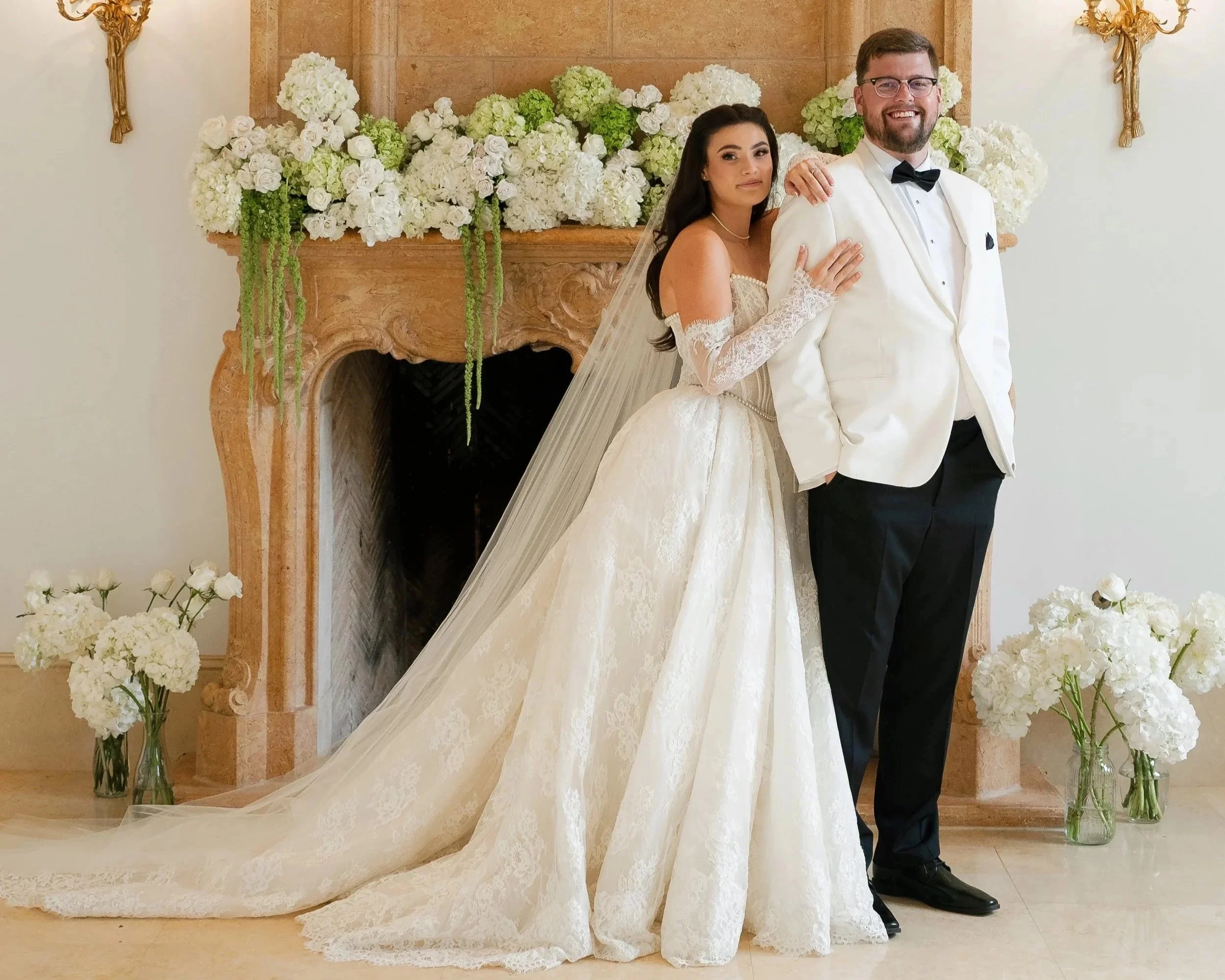 Bride and groom standing together in front of a decorated fireplace with white and green floral arrangements, the bride in a traditional white gown with lace sleeves and veil, and the groom in a white tuxedo jacket with black bow tie and black pants, smiling.