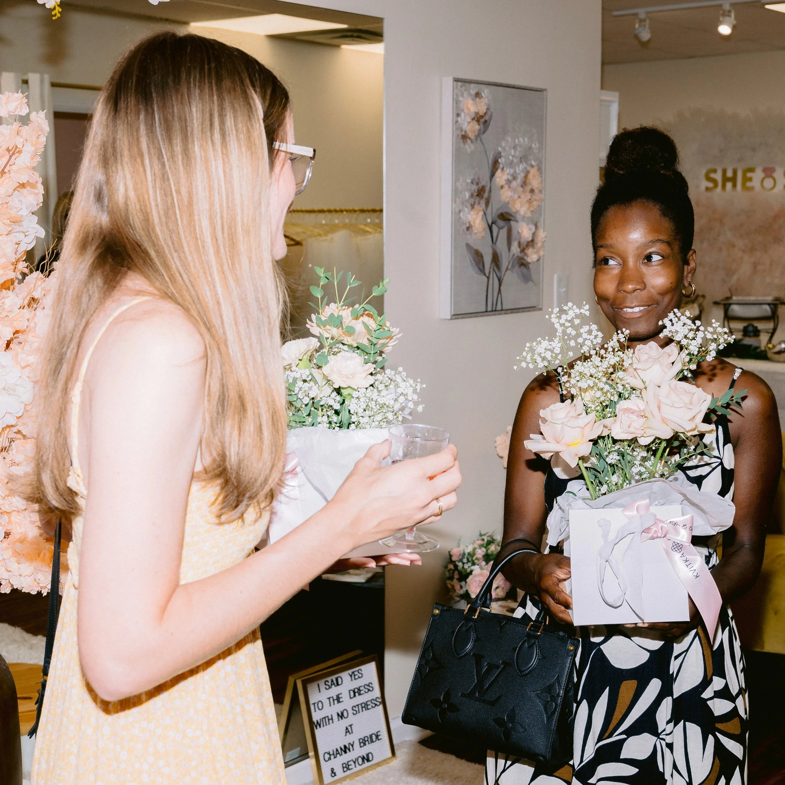 Two women standing and engaging in conversation indoors, one holding a bouquet of flowers, and a framed floral artwork on the wall behind them.