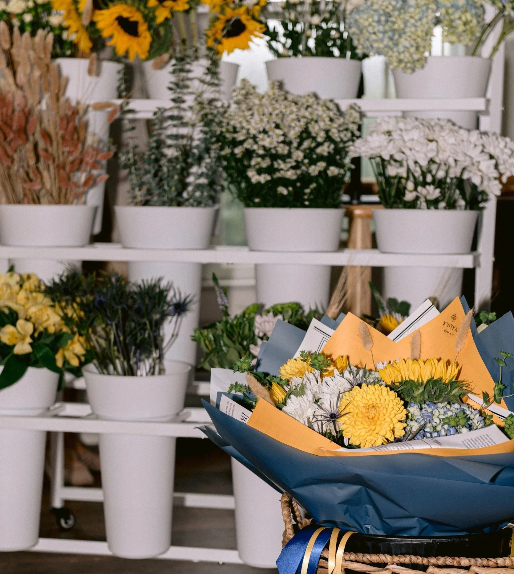 Various potted flowers on display in a flower shop, including sunflowers, daisies, and other blooms arranged in white pots and wrapping paper.