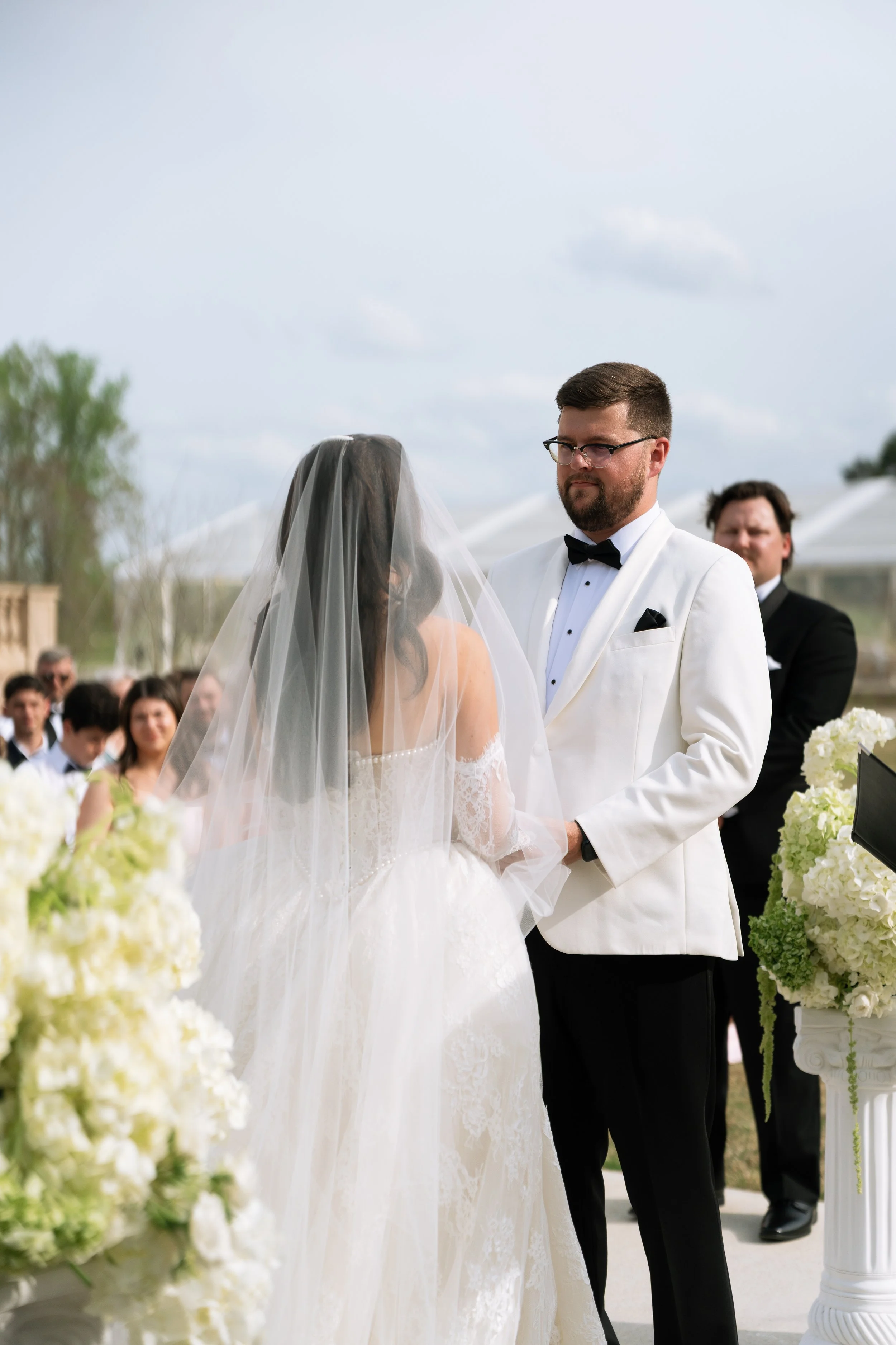 Bride and groom exchanging vows outdoors at a wedding ceremony, with guests in the background.