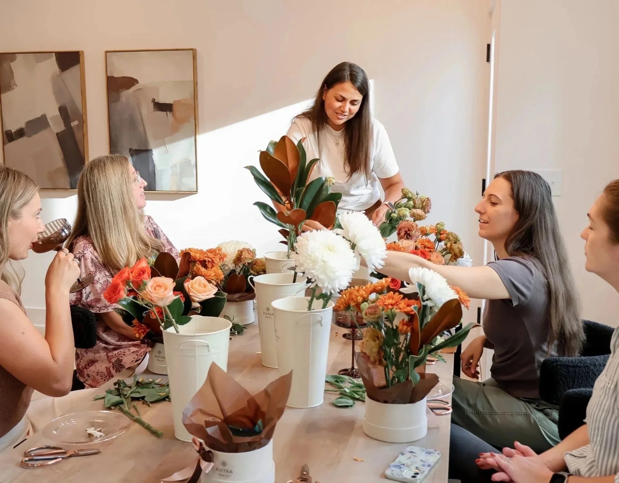Five women sitting around a table with flower arrangements and gardening supplies, one woman arranging flowers while the others watch and smile.