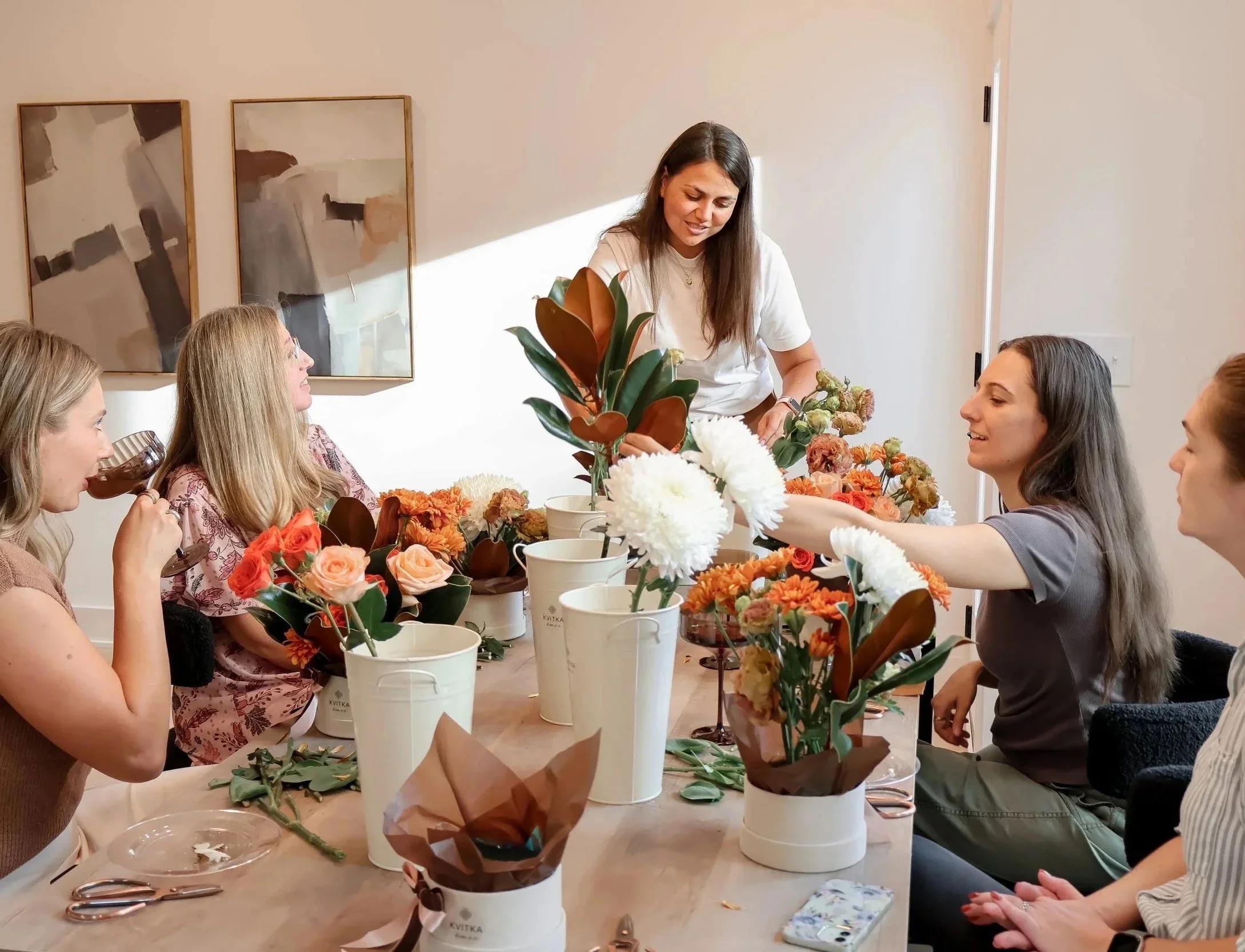 A group of women sitting at a table with flowers, participating in a flower arranging class, with a woman instructing.