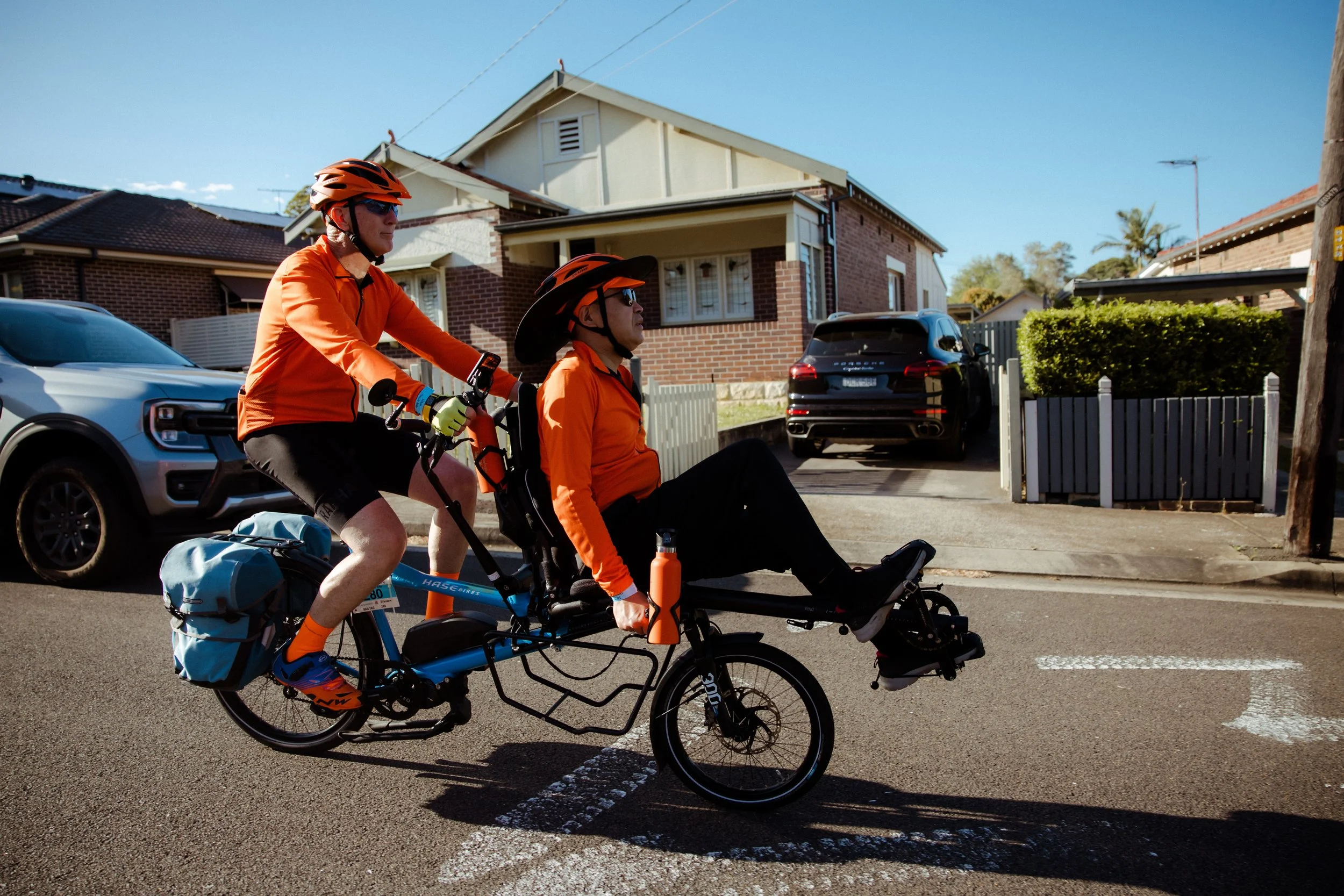 Two riders in orange on a semi-recumbent tandem riding through a Sydney street